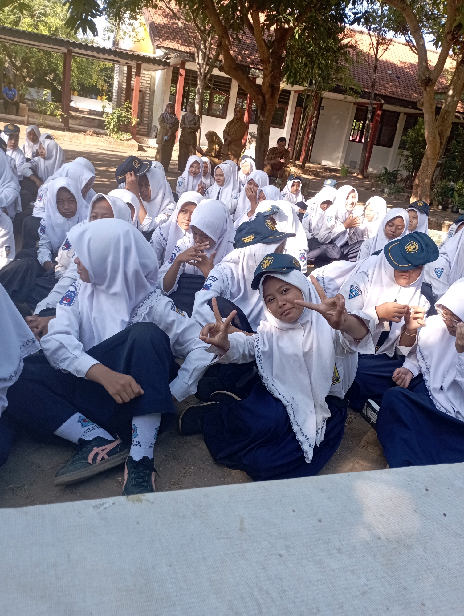 In Banyuurip, Indonesia, a group of approximately 20 female students in white uniforms and hijabs are sitting on the ground in an outdoor area, likely a schoolyard. The students are predominantly of Indonesian ethnicity and appear to be teenagers. The environment suggests a warm, sunny day, with trees providing dappled shade and a building with a tiled roof visible in the background. Some students are interacting, with one in the foreground making a peace sign with her fingers and another looking at her phone. The students' uniforms are white shirts with navy blue skirts or pants, and white hijabs. Some are wearing school hats. The ground is dirt and gravel. The overall atmosphere seems casual and relaxed, possibly during a break or an informal gathering.