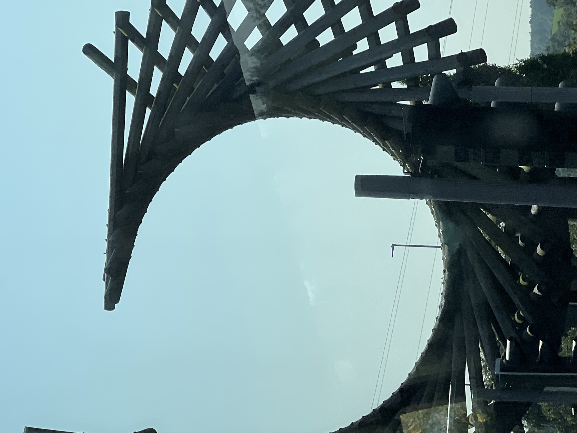 The image shows a large, dark, curved wooden structure, possibly part of a playground or art installation. The structure is made of numerous wooden beams arranged in a spiral or helical pattern, creating a series of open compartments. The sky is a pale blue, with a faint, wispy cloud. In the background, there are power lines and a lamppost. The lower portion of the image reveals more of the structure, including dark vertical supports and some smaller cylindrical elements with yellow caps. Trees and a green, hilly landscape are visible at the bottom edge of the frame. Based on available information, this structure is located in Letterkenny, Ireland, and is part of a skatepark. There are no people visible in the image.