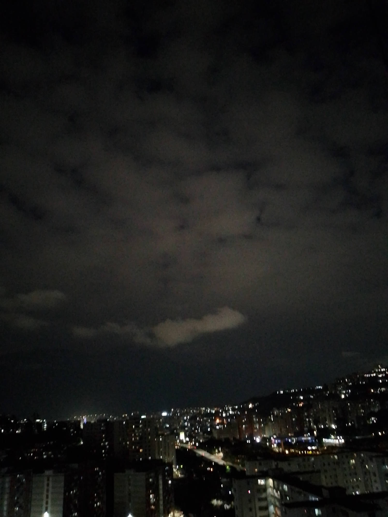 The image shows a night scene over a densely populated area, likely a city, with buildings and streetlights visible. The sky is dark with cloud cover. The presence of urban structures and lights indicates an urban setting, possibly in Caracas, Venezuela, given the query's context. The image does not reveal any people or specific actions. The scene's focus is on the cityscape at night under a cloudy sky.