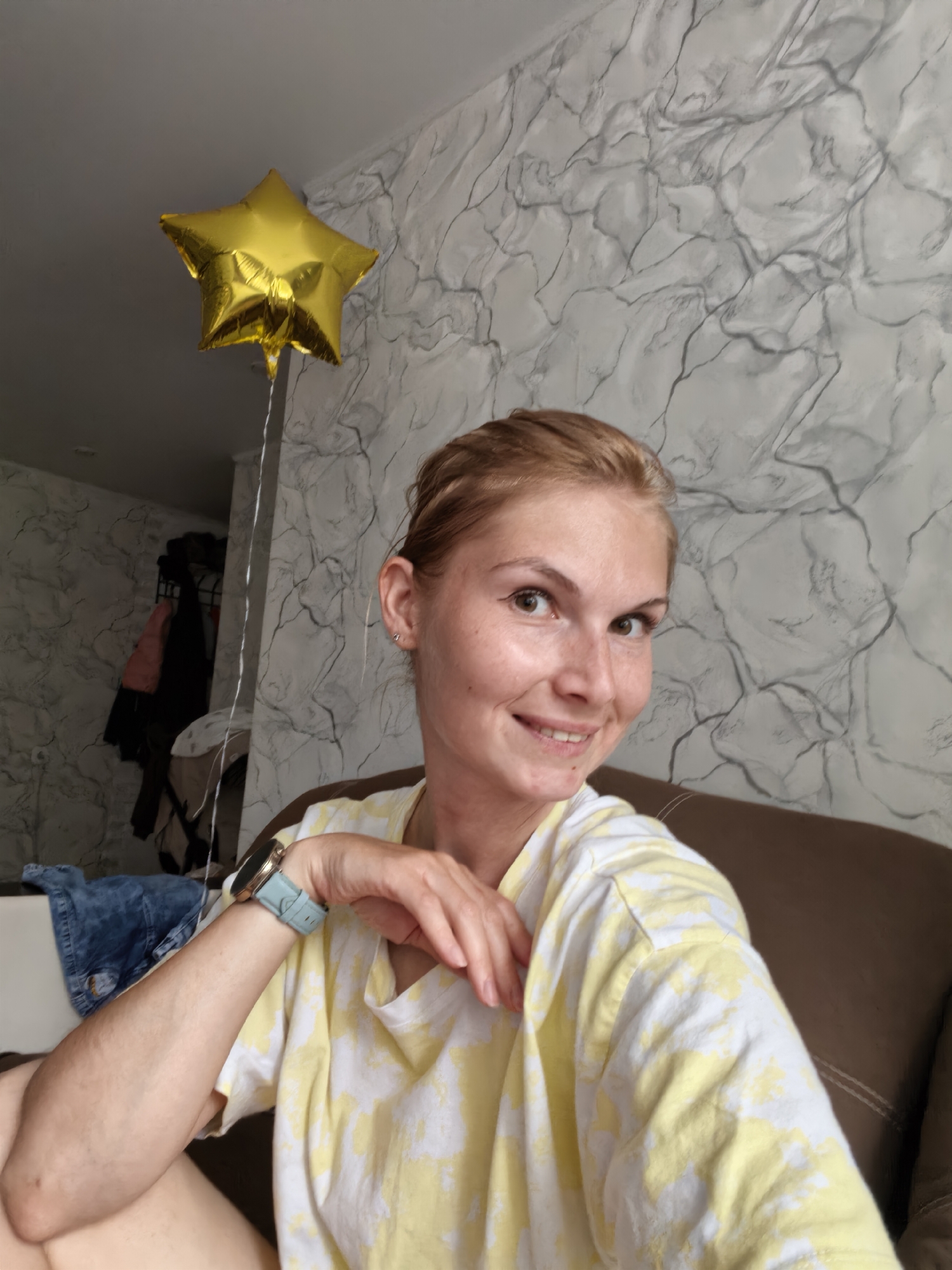 A woman is captured in a selfie from the chest up, seated on a brown couch. She has blonde hair pulled back and is smiling at the camera. She is wearing a yellow and white tie-dye t-shirt and a light blue wristwatch. To her left, a golden star-shaped balloon is visible, suspended by a string. In the background, a wall with a marbled grey and white pattern is depicted. To the left of the frame, a portion of a closet with hanging clothes and a pair of denim jeans is visible. The image does not provide any information or context that can be used to determine its location in the city of Sterlitamak, Russia.