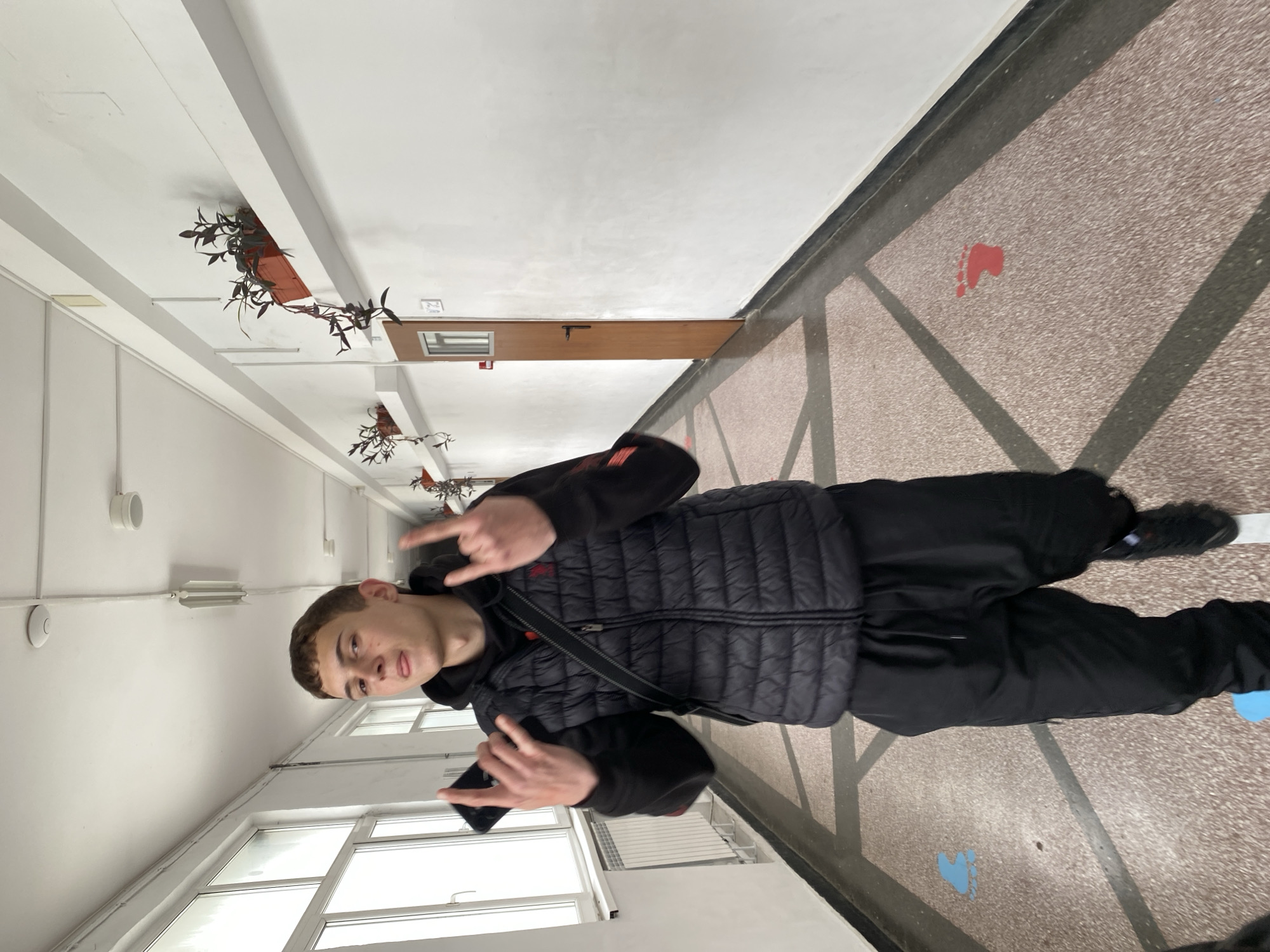 A teenage boy wearing a black puffer vest and black pants stands in a hallway. He's making a "rock on" hand gesture with both hands. The hallway has a speckled floor pattern and a white ceiling. There are plants in hanging containers on the ceiling. Footprint symbols are painted on the floor. The hallway is located in Varna, Bulgaria.