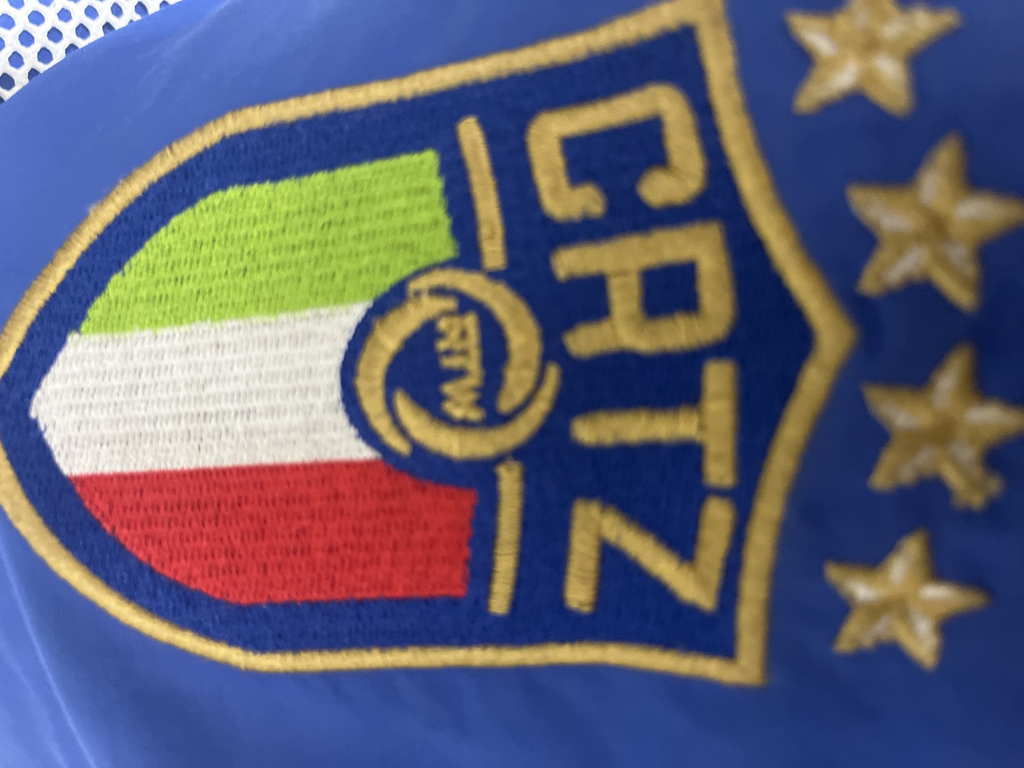 The image shows a close-up of a patch or emblem sewn onto a blue fabric. The patch is shaped like a shield and contains the colors of the Italian flag (green, white, and red) arranged horizontally. The letters "CRTZ" are vertically displayed in gold on the right side of the shield. A gold circular design is placed above the Italian flag stripes. Three gold stars are aligned vertically to the right of the shield. The scene does not provide any information about the location or the city of Yerevan, Armenia.