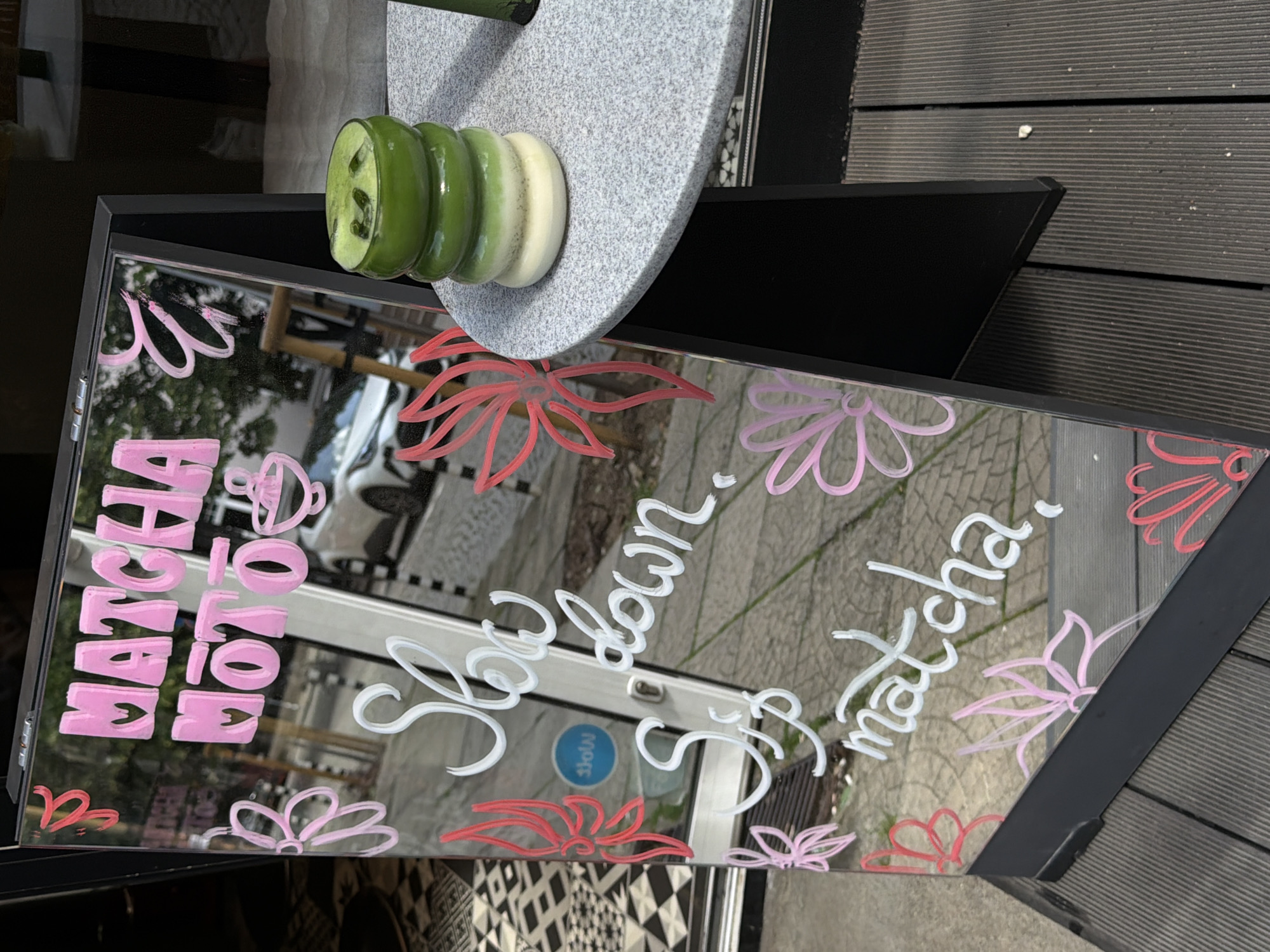 This image shows a close-up of a mirror with text and drawings on it, positioned outdoors. The mirror reflects a tiled or patterned floor and a street. The text on the mirror is in pink and white script, reading "MATCHA HOT & COLD" and "Slow down Sip matcha". There are also hand-drawn pink and red flowers on the mirror. The scene appears to be in a cafe or a similar establishment, possibly outdoors given the wooden decking visible in the background. A stack of green and white layered dessert is visible on a grey stone surface above the mirror, hinting at the cafe's offerings. The lighting suggests it is daytime. The overall aesthetic is vibrant and inviting, with a focus on promoting matcha products. The mirror is framed in black and attached to a black structure, possibly a display stand. The reflected scene includes a blurred view of a car and a blue circular sign with the word "slow" written on it.
