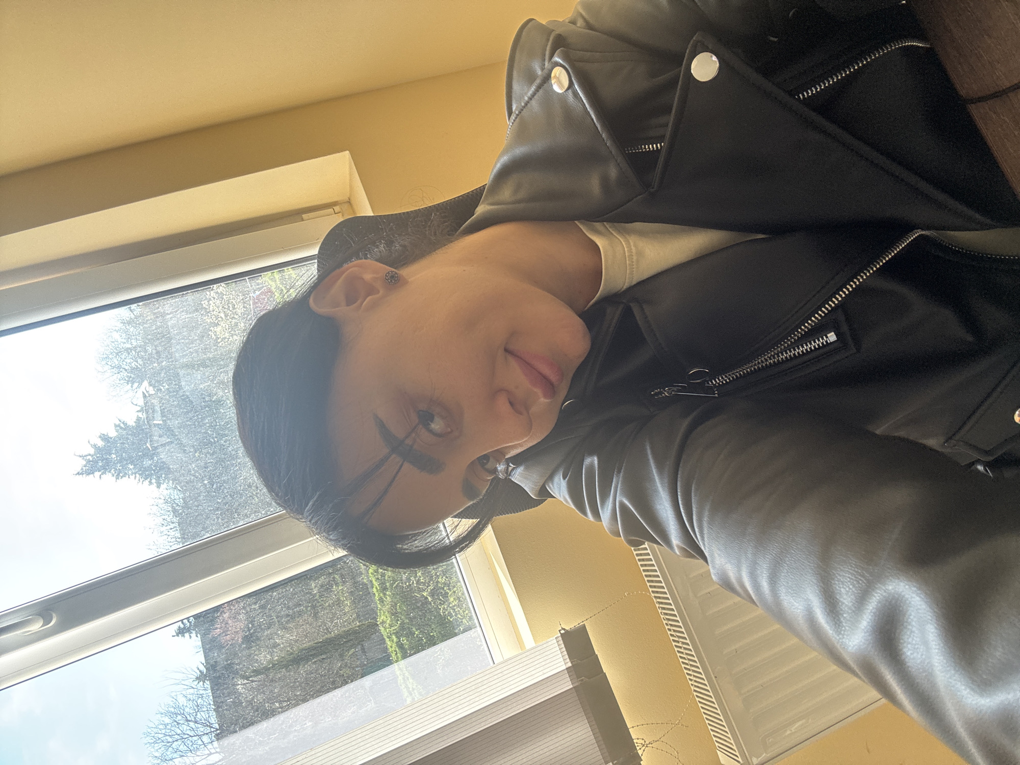 A person with dark hair, wearing a black leather jacket and a light-colored shirt, is positioned indoors near a window. The window offers a view of trees and buildings. The location is estimated to be in Tbilisi, Georgia. Blinds are visible within the room. The person appears to be taking a selfie.