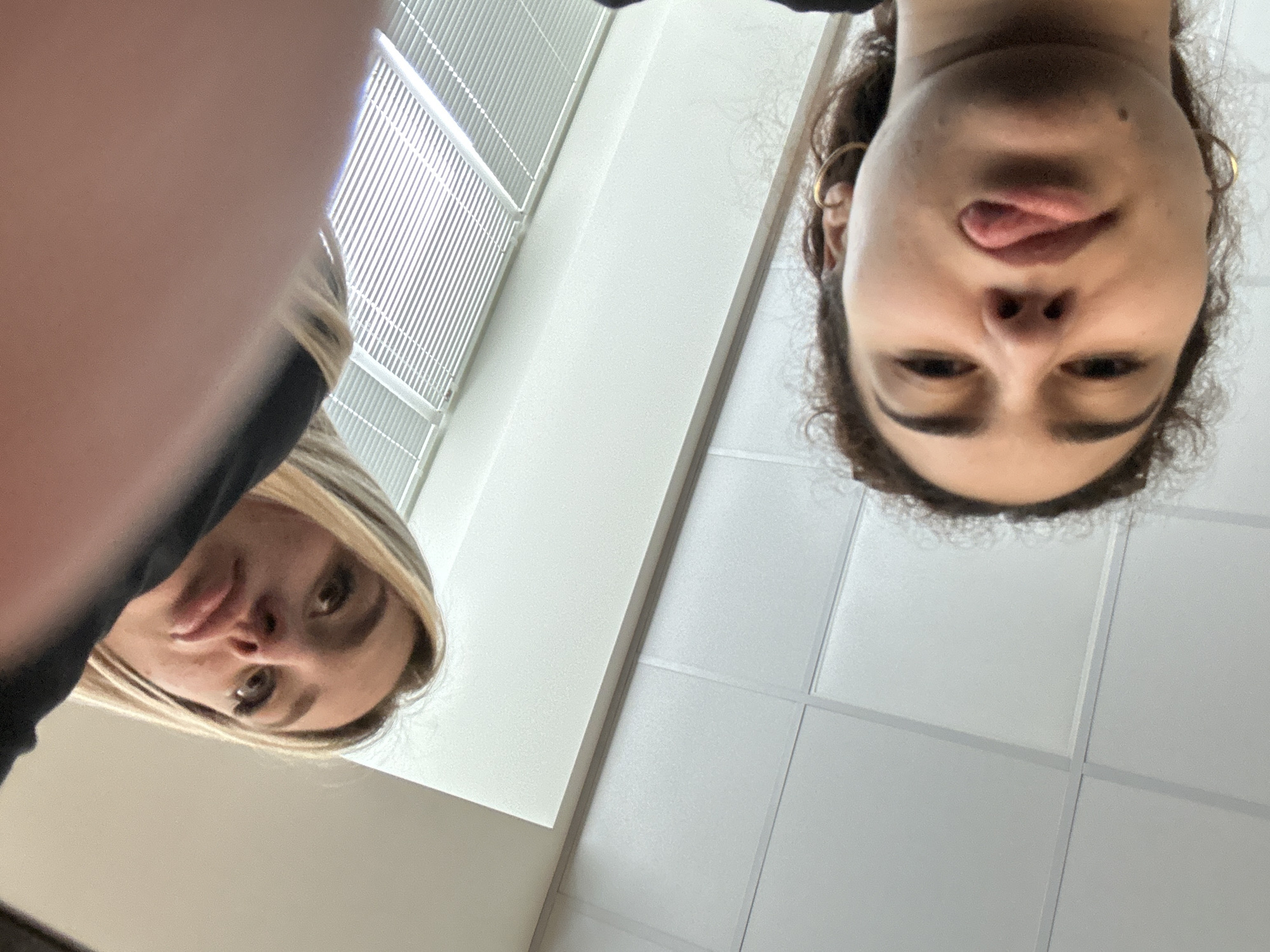 Two women are looking downward into the camera. One has blonde hair and is partially obscured by a dark garment in the left of the frame. The other woman has dark, curly hair and is sticking her tongue out slightly. In the background, there are white blinds and a white ceiling with a grid pattern. The image is taken from a low angle. There is no visible information to determine the location in Burgas, Bulgaria.