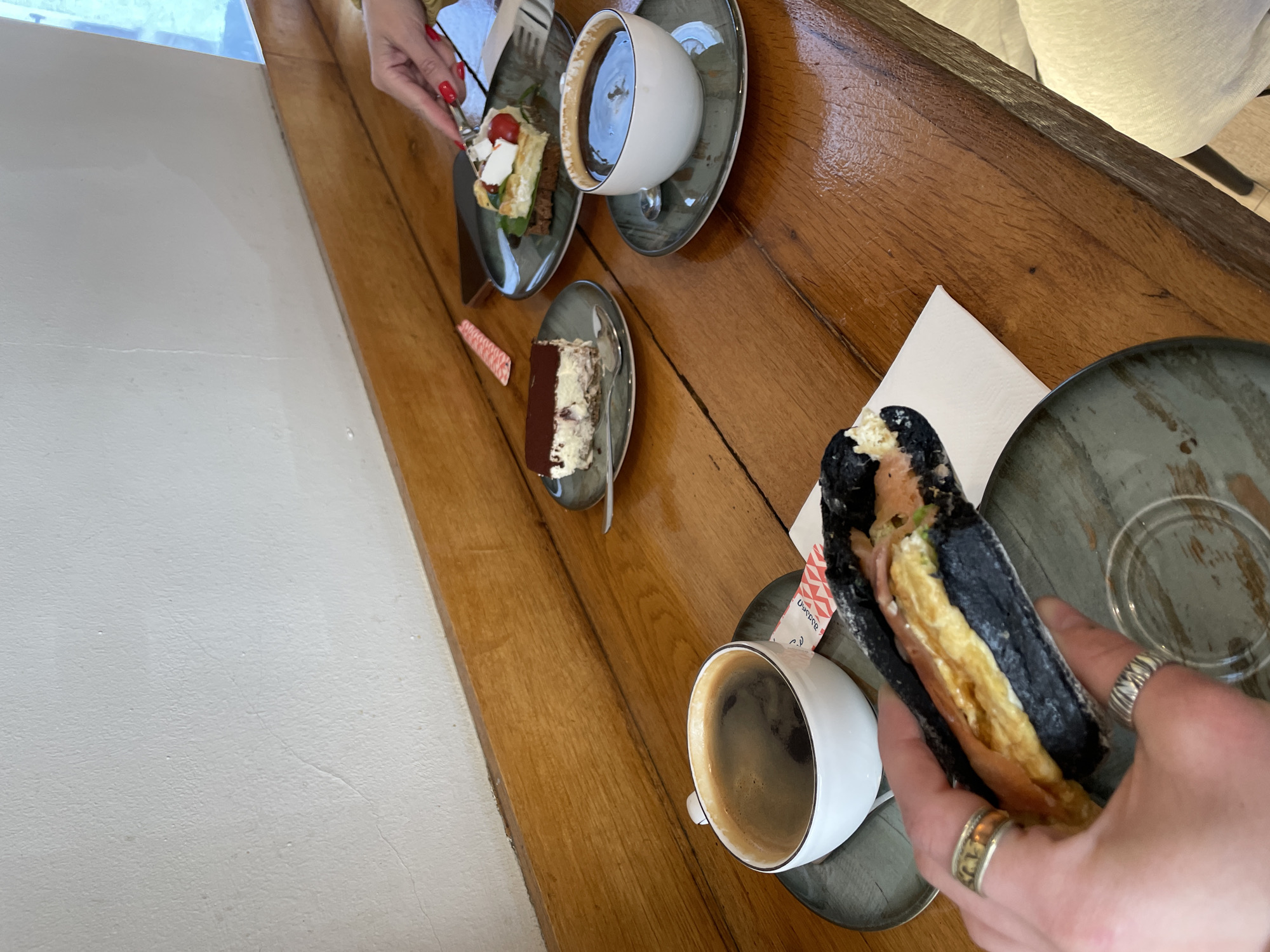 The image shows a wooden table in what appears to be a cafe. On the table are several plates with dessert items including cake and pastries. Two cups of coffee are present. One person is holding a black sandwich with filling. Another person is using a fork to eat a dessert. The location is likely Tbilisi, Georgia based on a sugar packet with Georgian script near the sandwich. The scene depicts individuals enjoying desserts and coffee at a cafe.