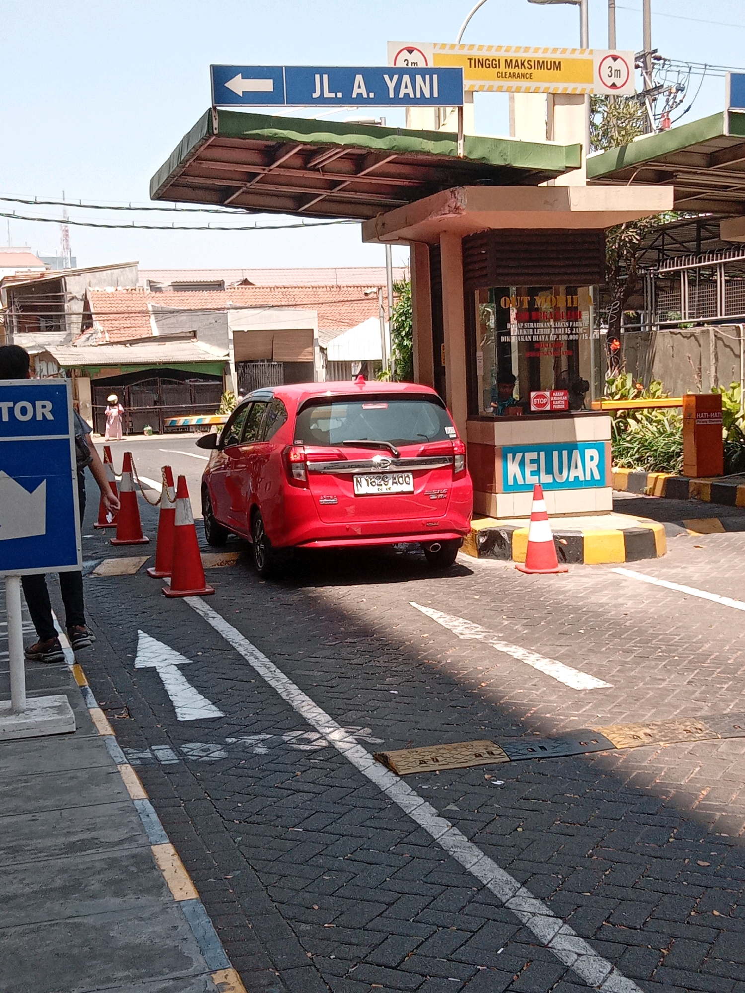 The image depicts an outdoor scene, likely a parking lot or entrance/exit area, in Surabaya, Indonesia. In the foreground, a red car is positioned on a paved surface. To the left, a blue sign with white text and a white arrow pointing left is visible, along with a person standing next to it. Orange traffic cones are placed in a line on the road. Behind the cones, a building structure with a "KELUAR" (Exit) sign is present, with a speed bump on the road in front of it. Overhead, a blue directional sign reads "JL. A. YANI" with a white arrow pointing left, and another yellow sign indicates "TINGGI MAKSIMUM" (Maximum Height) with measurements of "2m" and "3m". The background shows buildings and a clear sky.