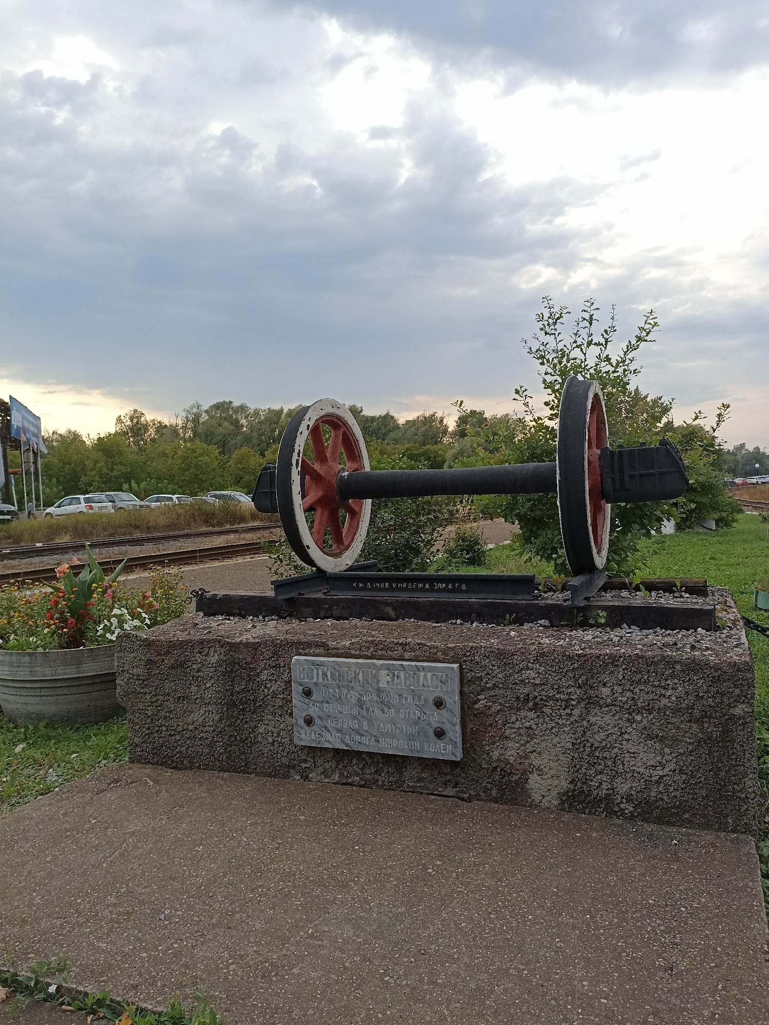 The image depicts an outdoor scene, likely a monument, in Votkinsk, Russia. In the foreground, a stone base supports a historical railway wheelset. The wheelset, consisting of two large, red and white wheels connected by a black axle, is mounted on a section of railway track. A metal plaque is affixed to the stone base. The plaque contains text in Russian, indicating a date and a reference to a railway opening. Behind the monument, railway tracks stretch into the distance, bordered by trees and vegetation. Several cars are parked near the tracks, and a cloudy sky forms the backdrop. A large concrete planter with flowers is visible to the left of the monument. The overall impression is of a historical marker or memorial located near a railway station.