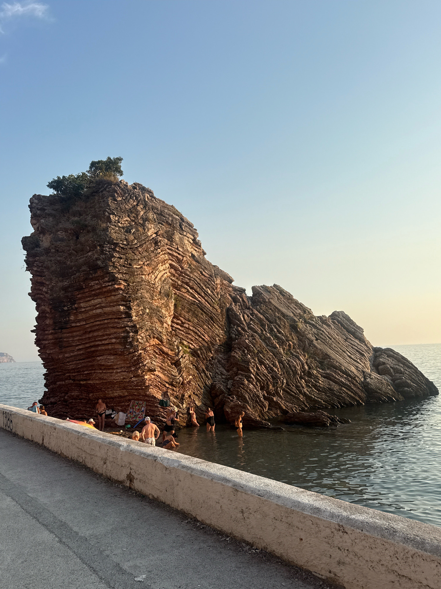 In Kamenovo, Montenegro, a large, stratified rock formation protrudes from the calm sea near a paved walkway. The rock, with its distinct horizontal layering and a small clump of trees on its summit, dominates the central and right portions of the image. The water surrounding the rock is clear and gently ripples. On the left side of the walkway, a group of approximately ten individuals are present. Some are seated on towels near the rock, while others are wading or standing in the shallow water. The sky above is clear with a pale gradient from blue to a lighter hue towards the horizon, suggesting it is either early morning or late afternoon. A white concrete barrier runs along the edge of the walkway, separating it from the water. Faint markings resembling the number "1888" are visible on the barrier. The overall scene depicts a serene coastal setting with people enjoying the water near a unique geological feature.