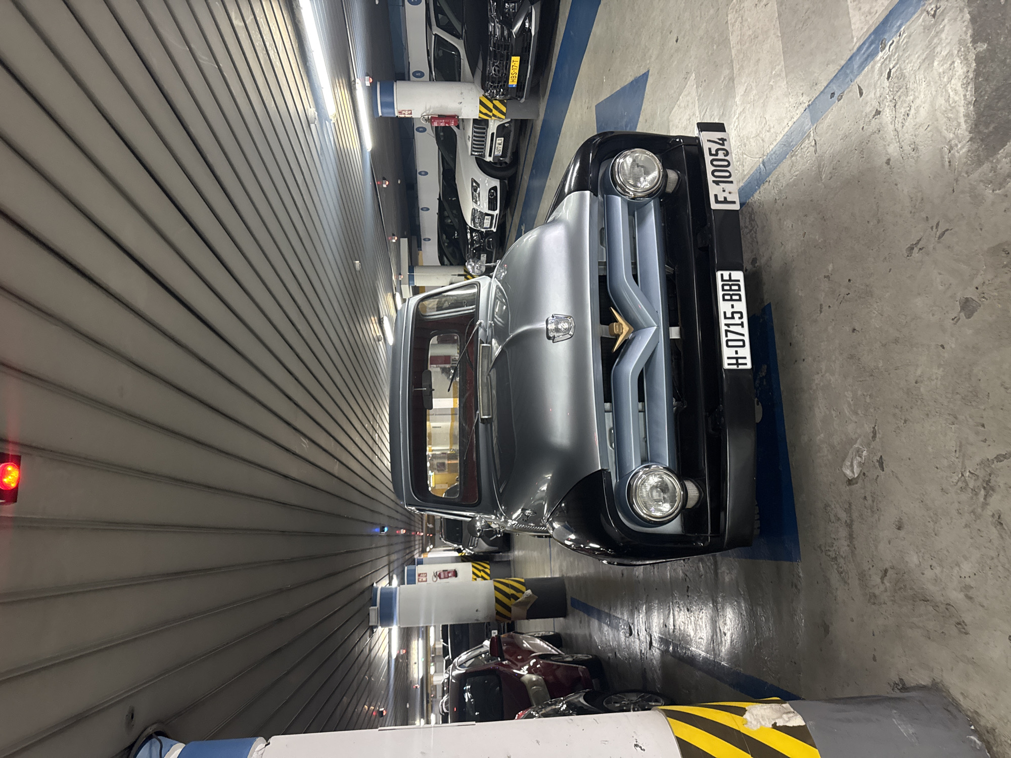 A two-tone vintage Ford F-series pickup truck, silver on top and black on the bottom, is parked in an indoor parking garage. The truck's front end, including its round headlights and chrome grille, is prominently displayed. The truck has two license plates: one reading "H-0715-BBF" and another reading "F-10054". Other vehicles, including a white Audi SUV and a black Hyundai SUV, are parked behind the Ford. The parking garage has a concrete floor, striped parking lines, and a corrugated metal ceiling with fluorescent lighting. In the background, there are parking garage columns with yellow and black hazard stripes. The scene takes place in Benidorm, Spain.
