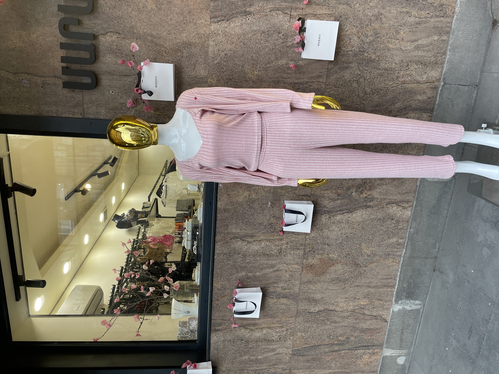 The image depicts a storefront display in Yerevan, Armenia. The store is named "Maravi" as indicated by signage and shopping bags. A mannequin with a gold head and white body is dressed in a pink striped ribbed knit set, consisting of a cardigan and pants. The mannequin is positioned against a brown marble-like wall. The display includes white shopping bags with black handles and the "Maravi" logo. Artificial pink flowers are scattered around the bags and the mannequin. The storefront window shows interior details, including other clothing items, lighting fixtures, and architectural features of the store. The sidewalk is visible on the right edge of the frame.