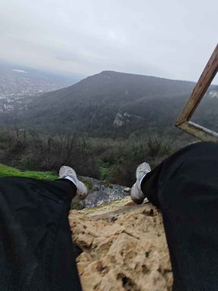 The image depicts a first-person perspective from a high vantage point overlooking the city of Shumen, Bulgaria, and the surrounding forested Shumen Plateau. In the foreground, the lower legs of an individual, dressed in black pants and light-colored sneakers, are extended over a rough, yellowish-brown rock surface, indicating placement at a cliff edge. Directly below and extending into the midground is a steep, densely forested slope, primarily consisting of bare or sparsely leaved deciduous trees, with patches of green vegetation. A prominent light-colored rock face is visible within the trees on the right midground. To the left, a sprawling urban area, identified as Shumen, is visible in the distance, with numerous buildings and structures. Beyond the city, a lighter-colored expanse, possibly an agricultural field or distant body of water, stretches towards the horizon. The background features additional undulating landforms under a uniformly overcast, light grey sky. A rusted, angular metallic structure, potentially part of a railing or platform, is partially visible in the upper right.