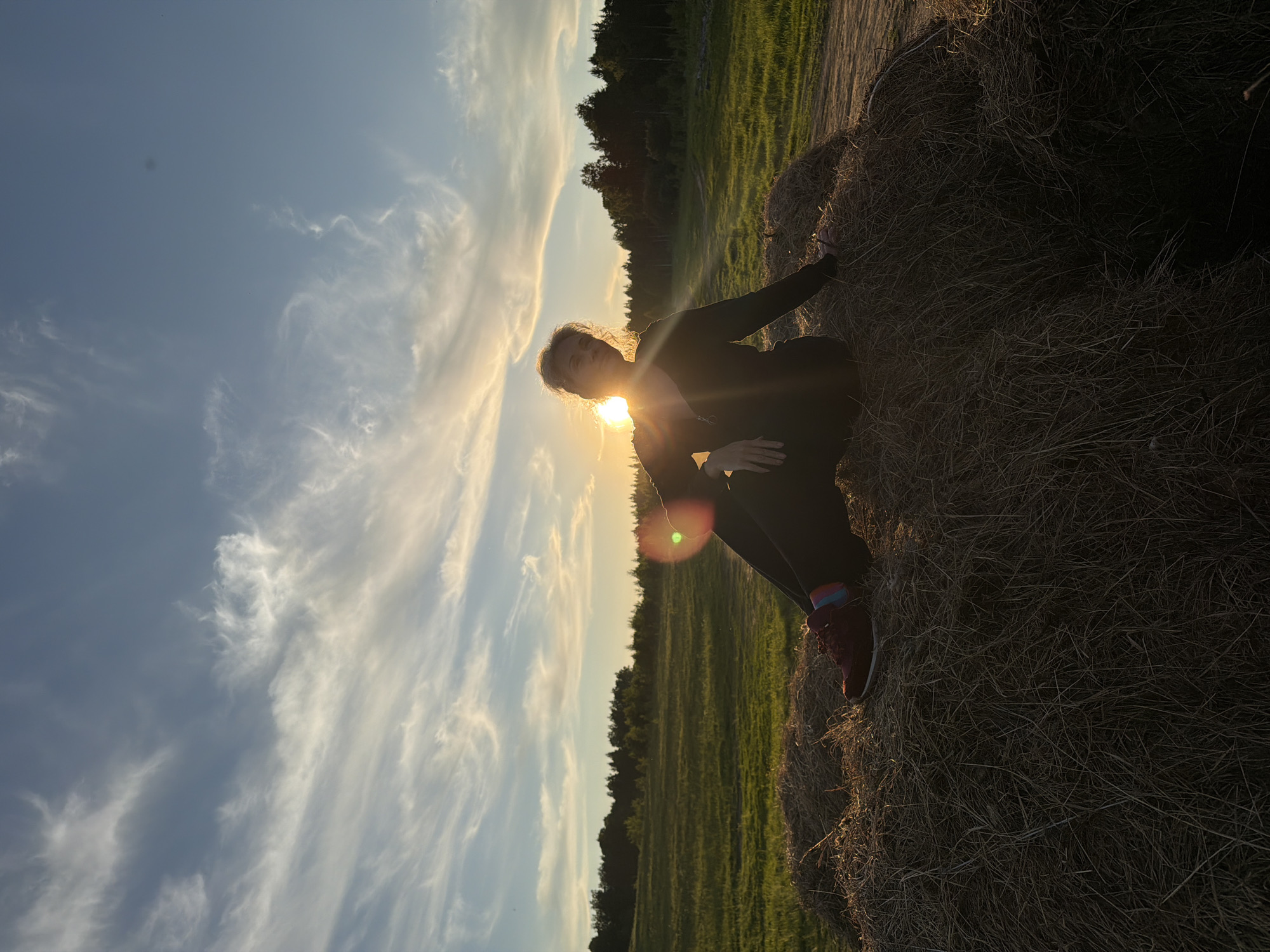 The image, rotated 90 degrees clockwise, captures a woman crouching beside a large round hay bale in a rural landscape during golden hour. The sun, likely setting or rising, is positioned directly behind her head and right shoulder, creating a strong backlighting effect. This illuminates her light-colored hair with a halo and silhouettes her figure, obscuring facial details.
The woman is dressed in dark clothing, possibly a dark jacket or hoodie and pants. She wears striking red or maroon athletic shoes with multicolored striped socks. Her left hand rests on the hay bale, while her right hand is near her lap. A prominent green circular lens flare is visible near her right leg due to the direct sunlight.
The setting includes a vast, flat green field stretching into the distance, bordered by a dark line of trees or a forest. The sky is a mix of clear blue and warmer hues near the sun, with scattered wispy clouds. The atmosphere is serene and natural, consistent with a countryside location like Ozernoye-1, Russia. There is no visible text in the image.