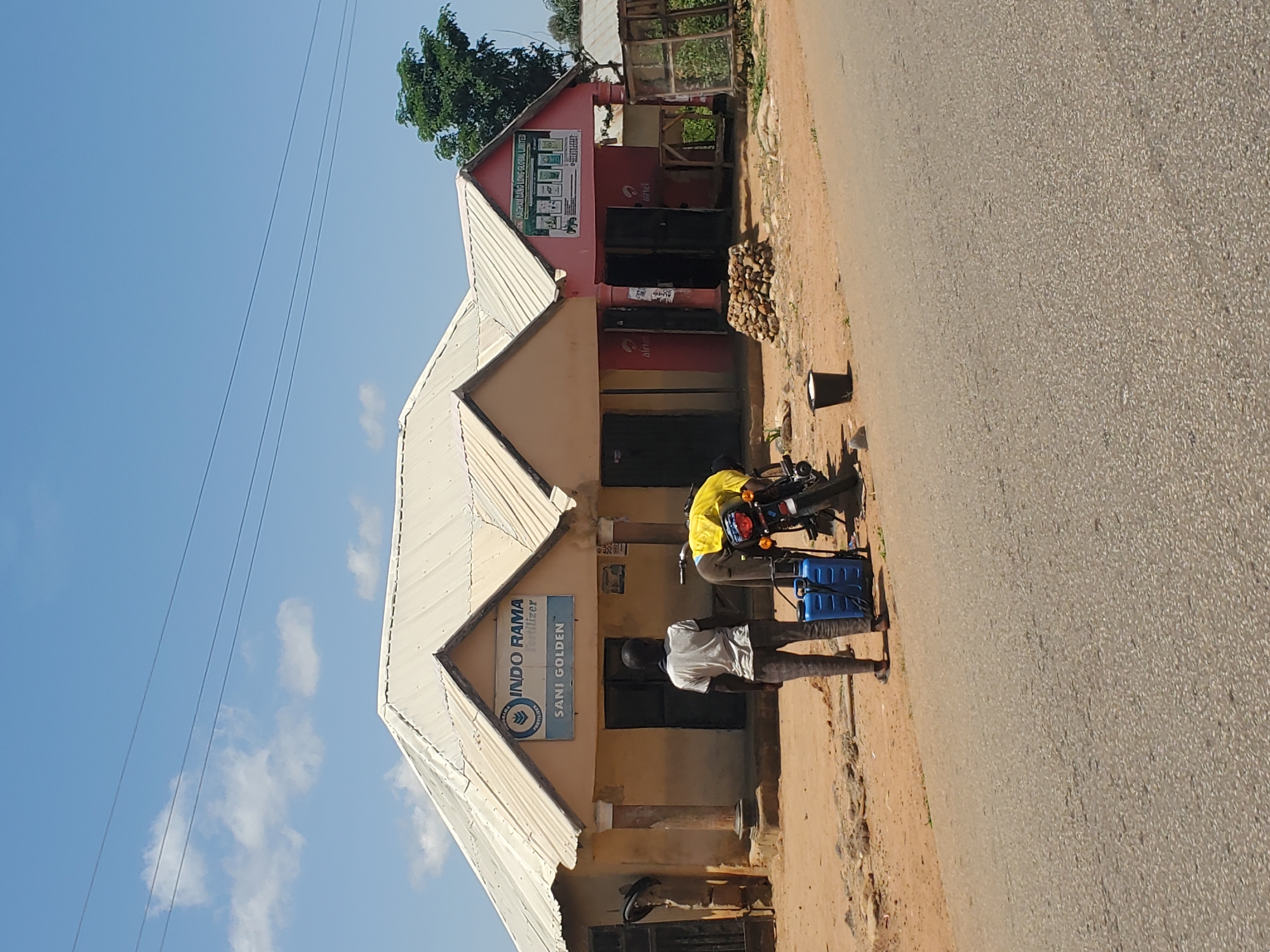 In Mangu, Nigeria, a sunny, outdoor scene unfolds with a focus on a commercial strip. Two buildings with white corrugated metal roofs line a dirt road. The left building, painted a light brown, features a prominent sign for "INDO RAMA Fertilizer SANI GOLDEN". Two pillars support its facade, and a dark doorway is visible. To the right, a pink building displays a green and white sign that reads "KAGRAN DANG LONG GLOBAL LIMITED", also with multiple product images. Red Airtel logos are present on both buildings, indicating telecom services.
In front of the buildings, a young man in a gray t-shirt and dark pants stands with his back to the camera, facing the "INDO RAMA" building. Beside him is a bright blue sprayer with a handle. A motorcycle, partially obscured, has a yellow cover draped over its rear. A black bucket sits near the motorcycle. The ground is dusty, with scattered debris and stones. The road in the foreground is paved, contrasting with the unpaved area in front of the shops. A mature green tree stands behind the pink building. The sky is a clear blue with a few wispy white clouds, and thin electrical wires are strung overhead.