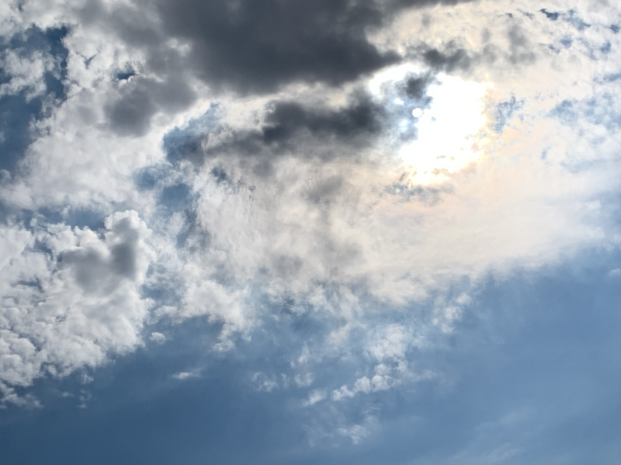 This is a wide-angle, eye-level, full-frame shot of a cloudy sky taken in Khazar Raion, Azerbaijan. The scene is dominated by a mix of white and dark gray clouds. The sun is visible as a bright, blown-out white circle peeking through a break in the darker clouds, casting a strong glare across the upper right quadrant of the image. The lower portion of the image transitions into a clear, light blue sky. There are no people, animals, or man-made structures visible.