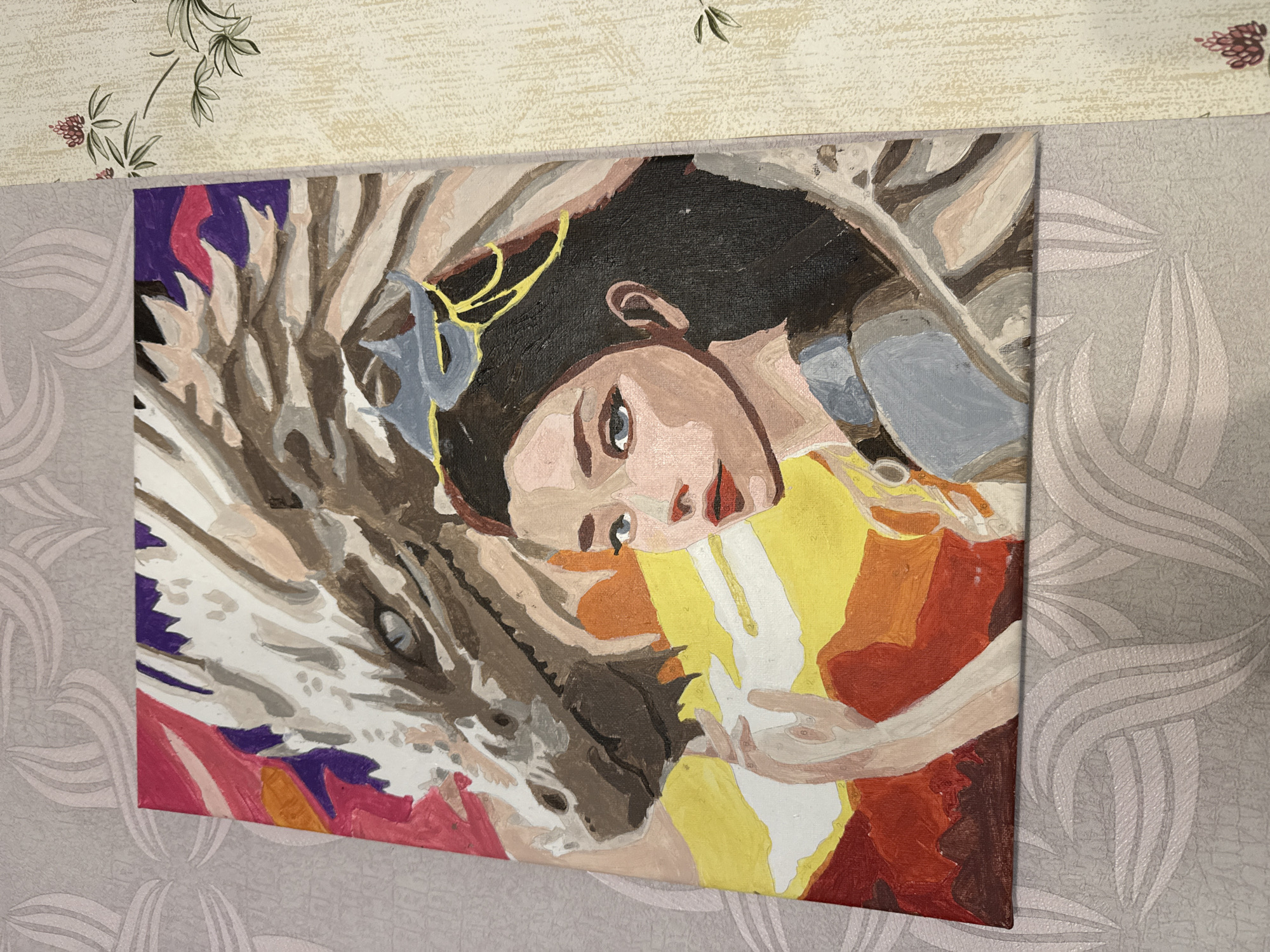 This is a close-up, top-down shot of an acrylic painting on a canvas, depicting a fantasy scene. The painting features a young woman with dark hair and blue eyes, wearing a blue and grey garment and a golden headpiece, positioned in the foreground. To her left, a large, dragon-like creature with white and brown scales and blue eyes is visible, with its snout close to the woman's face. The background is a vibrant blend of red, orange, yellow, pink, and purple, suggesting a fiery or dramatic setting. The canvas is resting on a surface with patterned wallpaper, one section a light grey with a large, abstract floral design, and another section a cream color with a smaller floral motif. The lighting is consistent, indicating an indoor setting with diffuse light.