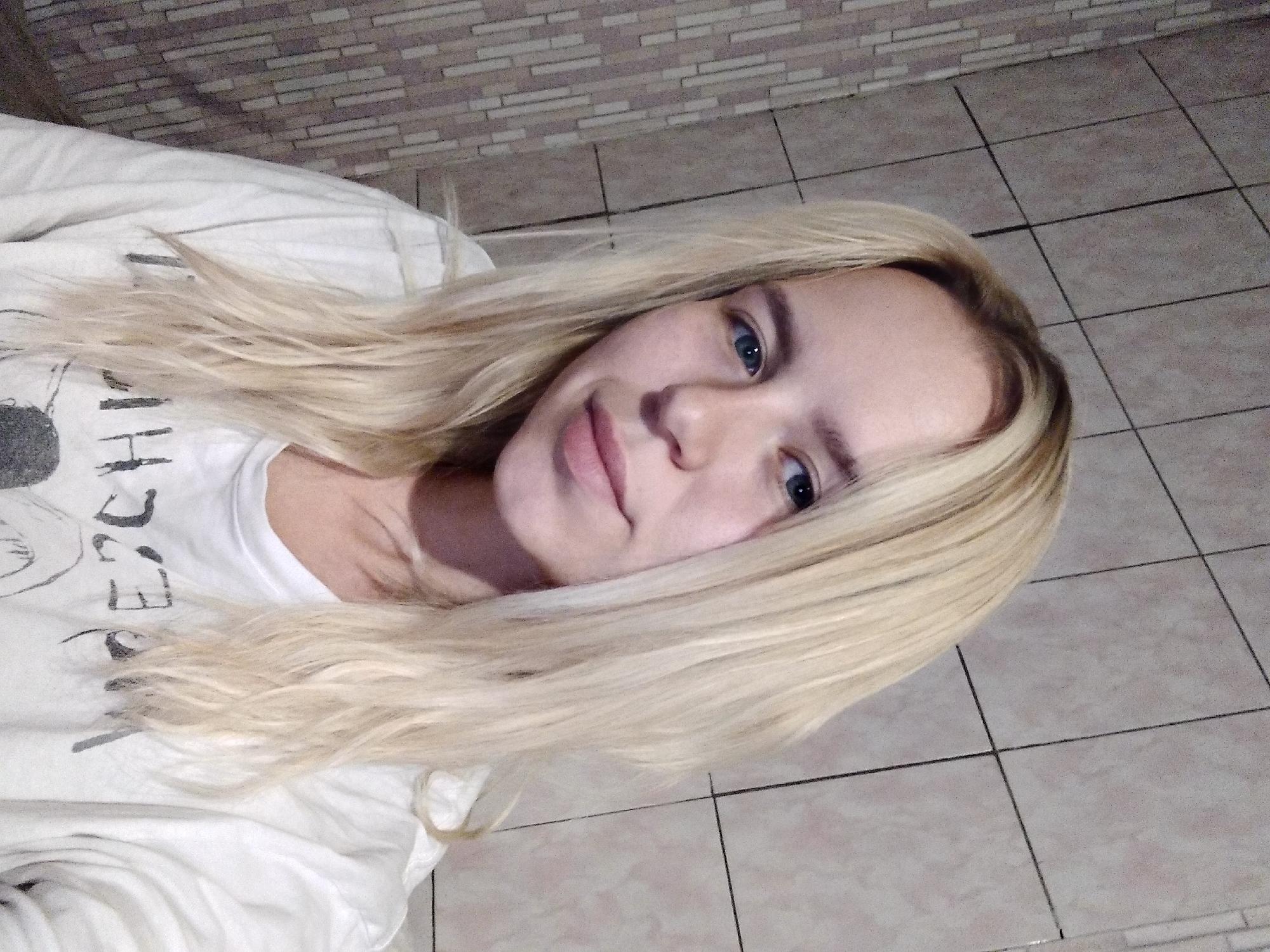 A young woman with blonde hair and blue eyes is taking a selfie in what appears to be a bathroom. She is wearing a white t-shirt with a graphic design on it. The background consists of tiled walls.