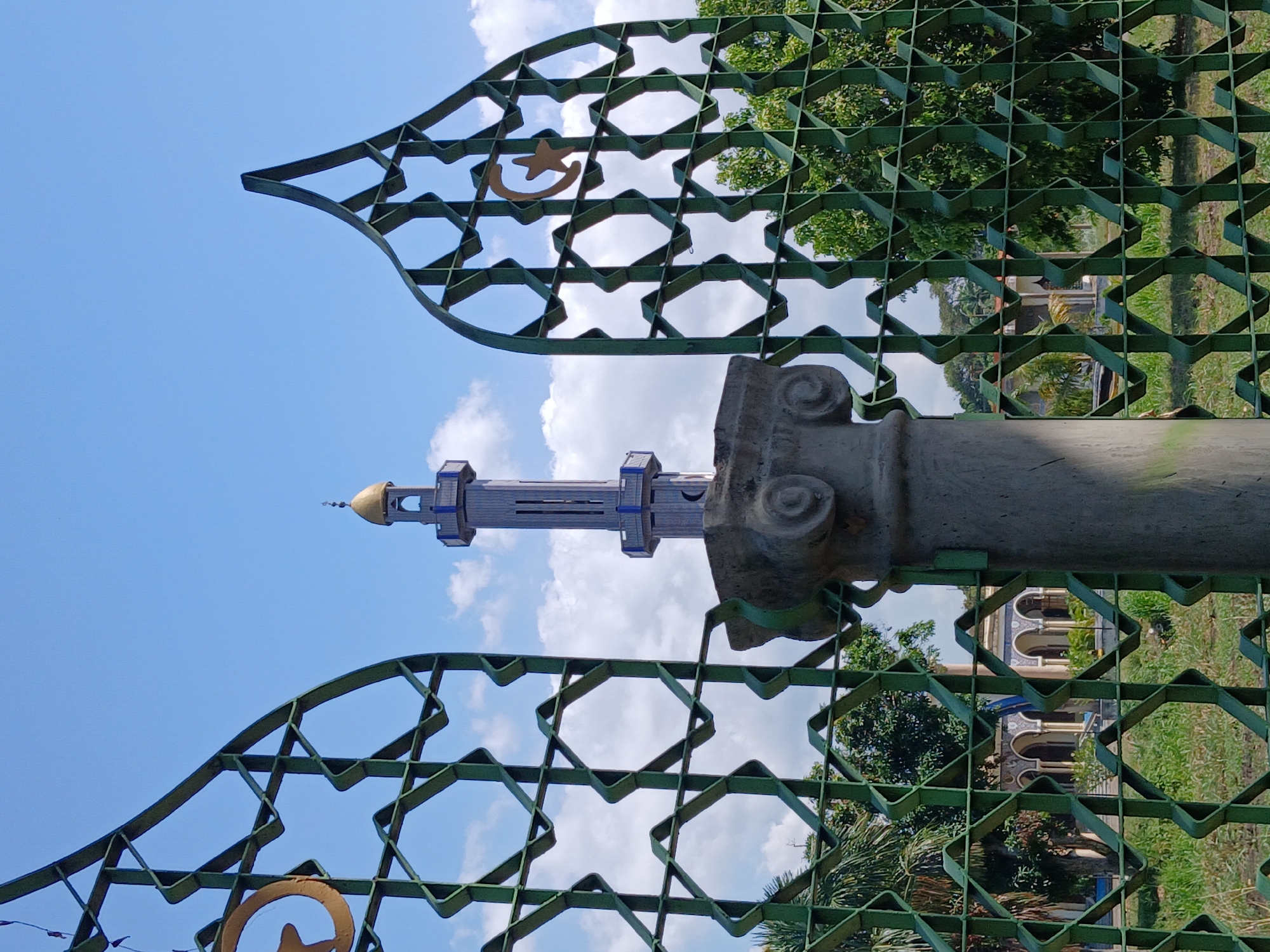 A vertical outdoor shot shows an elaborate, green metal gate with repeating geometric shapes and star and crescent symbols. Behind the gate is a gray, classical-style pillar and a mosque minaret with a golden dome against a sky with white clouds. The background includes trees, grass, and a glimpse of another building with arches. The image appears to be taken in San Felix, Venezuela.