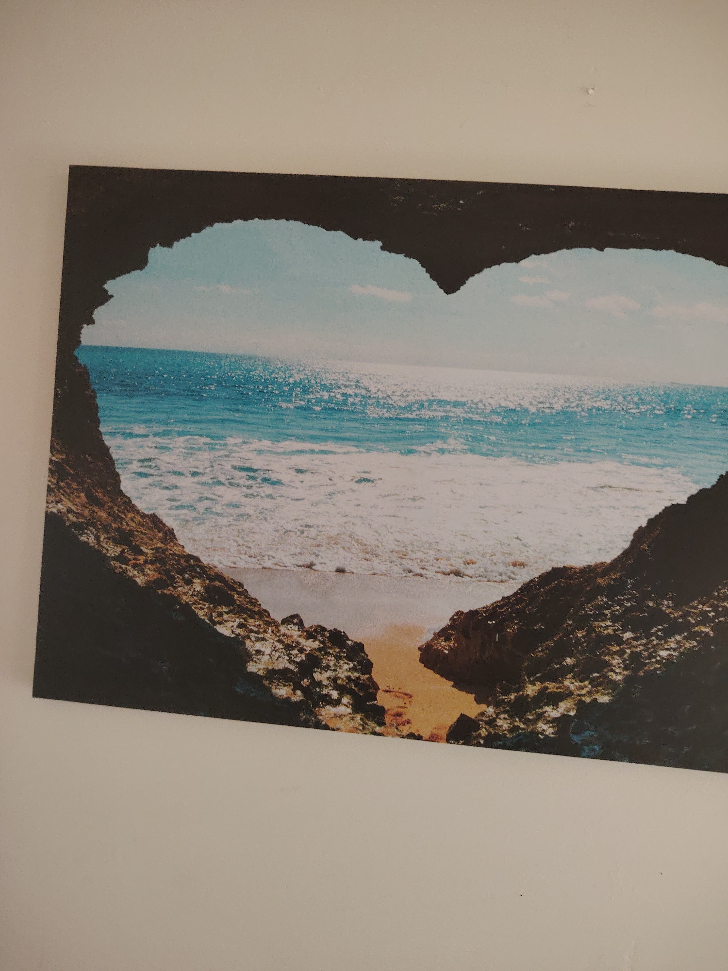 The media file displays a photograph of a rectangular canvas print, mounted slightly off-kilter on a plain, light-colored wall. The print itself features a picturesque coastal scene, artistically framed by a natural, heart-shaped opening in dark, rugged rock formations.
Through this heart-shaped archway, a vibrant ocean stretches into the distance under a bright, light blue sky with hints of faint clouds. The sea exhibits rich blue and turquoise hues, with numerous sparkling reflections of sunlight creating a shimmering effect across its surface. Below, foamy white waves gently break onto a narrow strip of sandy beach, which is a light brown color. The dark, textured rocks forming the heart shape appear to be part of a cave or a natural rock arch, providing a striking contrast to the luminous seascape within.
No people, animals, or specific activities are visible within the depicted scene, emphasizing the natural beauty and tranquility of the coastal environment. The bright sun reflections on the water suggest a clear, sunny day, likely mid-day. The overall impression conveyed by the print is one of serenity, natural wonder, and romance. There is no visible text within the artwork. The surrounding wall is a uniform off-white color, showing a small pinhole and minor speckles in different cropped sections.
