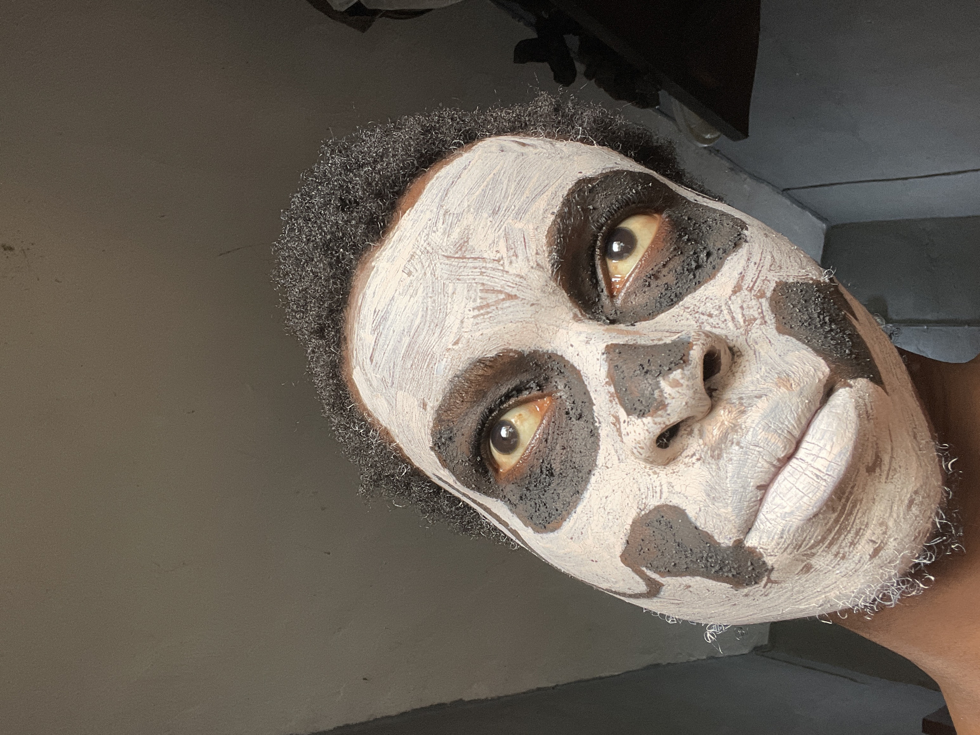 A close-up, angled portrait shows an individual, likely an adult male of African descent, with short, dark, curly hair. Their face is covered in a distinctive white and dark (possibly black or dark brown) face mask or paint. The white layer, applied with visible brushstrokes or finger marks, covers large areas of the forehead, cheeks, chin, and lips. Dark, textured patches of material are prominently placed around the eyes, forming thick borders, and also appear as irregular spots on the cheeks and around the nose.
The person's eyes are wide open, directly gazing at the camera, with the sclera exhibiting a slight yellowish tint. The lips are coated in the white mask, appearing dry and textured. The head is tilted sharply to the left, presenting an unconventional, almost abstract, composition.
The background is simple and dimly lit, consisting of plain, light gray walls. Darker, out-of-focus elements are visible in the upper corners, suggesting an indoor setting, possibly a room with minimal decor. The activity appears to be posing for a photograph, likely showcasing the face application which could be for artistic expression, a costume, or a skincare routine. No text is visible in the image. The scene is indoors, consistent with the given location context of Onitsha, Nigeria.