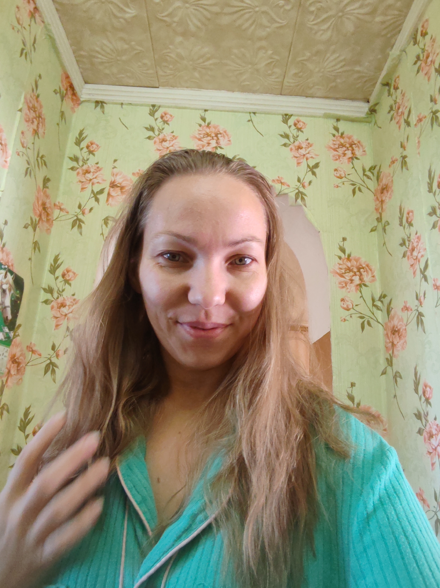 A medium shot portrait of a woman with long, light brown hair, looking at the camera with a slight smile. She is wearing a teal-colored garment with a white and pink trim at the collar. Her left hand is raised, fingers slightly splayed, near her hair. The background consists of light green wallpaper with a floral pattern of pink and peach flowers and green leaves. Above the wallpaper, a white trim separates it from a beige, textured ceiling with a repeating embossed pattern. To the left, partially visible on the wall, is a framed picture of a horse. The lighting suggests an indoor environment, possibly a home. The image is taken from a slightly low angle. There are no discernible elements that indicate the specific location within the city of Чухлэм, Russia.