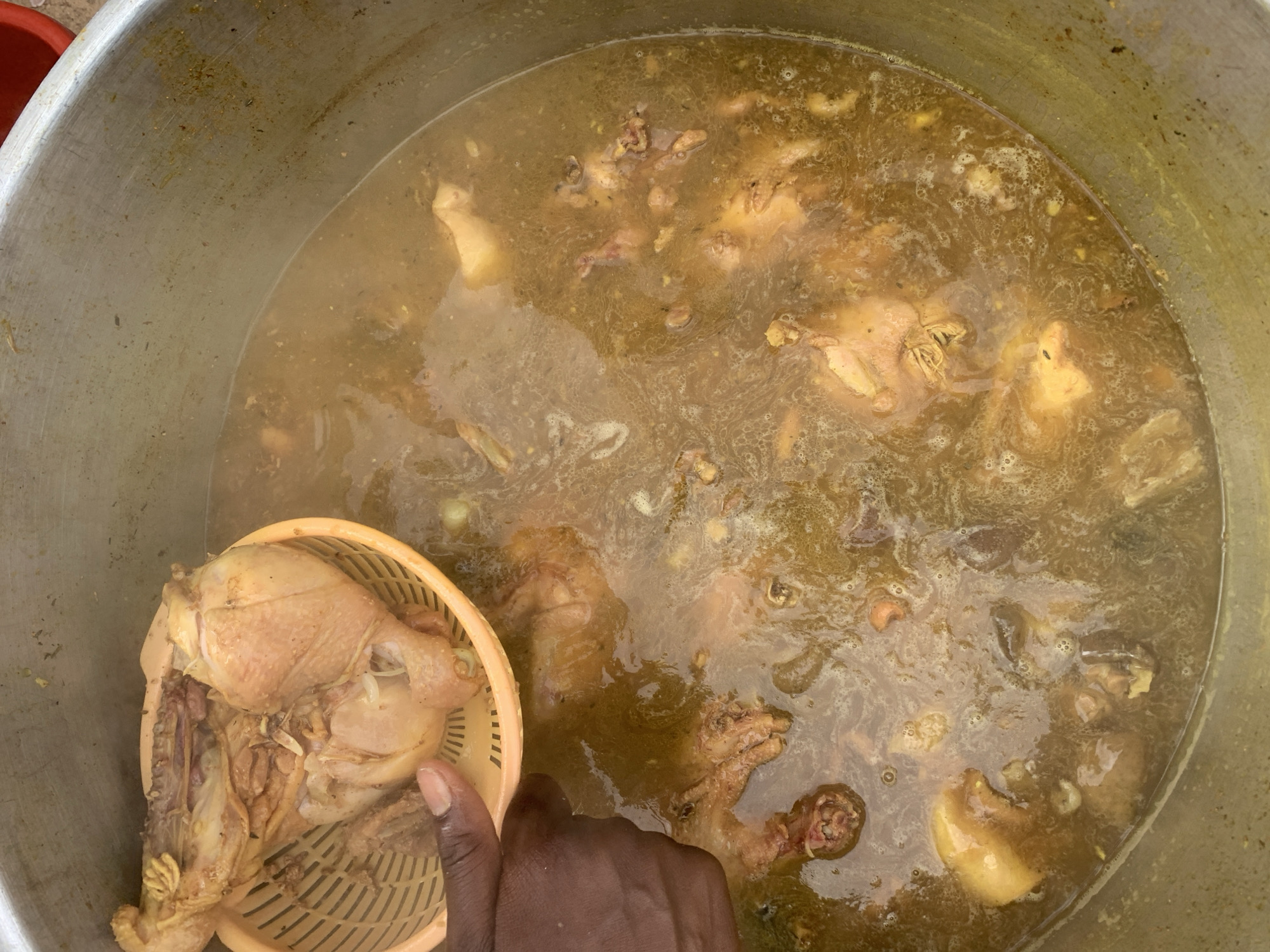 The image captures an overhead view of a large-scale cooking operation in New Stadium, Nigeria. A dark-skinned adult hand, positioned on the left, holds a light orange plastic colander containing several large pieces of cooked, pale-yellow chicken with visible bones and skin. This colander is suspended directly over a very large, utilitarian, metallic cooking pot, which dominates the central and lower portions of the frame. The pot contains a simmering, murky, yellowish-brown liquid stew, within which numerous pieces of chicken and other ingredients are partially submerged. Small bubbles are visible on the liquid's surface, indicating heat. The interior rim and upper wall of the pot show signs of use and minor staining. In the upper right corner, a small, out-of-focus section of a red object is visible, hinting at the broader environment.