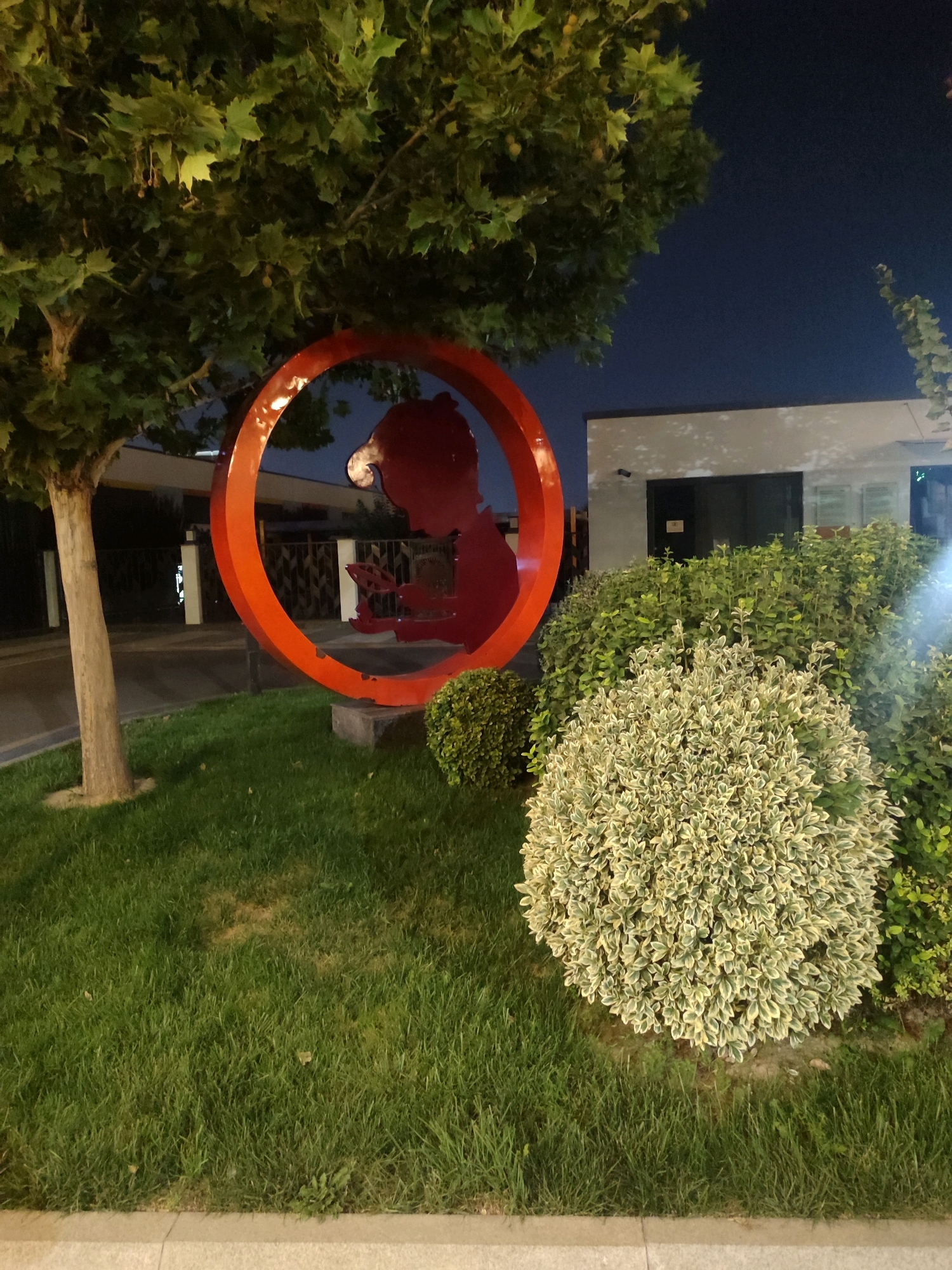 An outdoor shot at night captures a red circular sculpture featuring a silhouette of a person reading a book against a dark blue sky. The sculpture is positioned on a patch of green grass, with a large tree to its left and a manicured bush to its right. In the background, a modern building with white walls and dark windows is visible, suggesting an urban setting. The image is likely taken in the city of Tashkent, Uzbekistan, given the architectural style and landscaping.