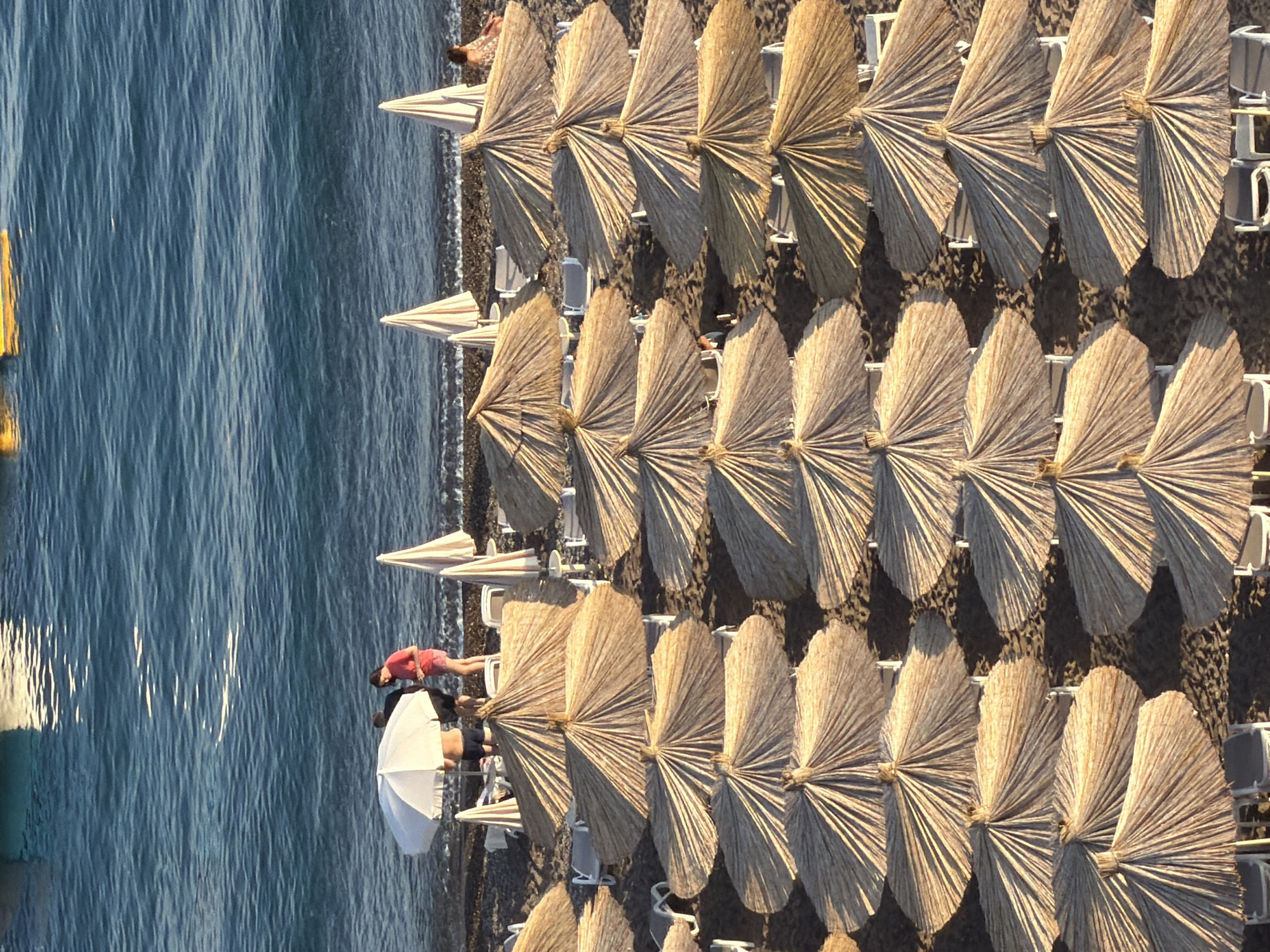 An aerial view captures a vibrant beach scene in Boreti, Montenegro, under clear skies. The tranquil, blue-green sea occupies the left side of the frame, with gentle ripples on its surface and a yellow float partially visible near the shoreline.
To the right, the dark pebble or sand beach is neatly organized with multiple rows of beach umbrellas and sunbeds. Many umbrellas are closed, forming pointed conical shapes, while others are open, fanning out in a distinctive straw-like or wicker-like pattern. The warm, directional sunlight casts long shadows from these structures, suggesting either early morning or late afternoon.
Several individuals are visible enjoying the leisurely beach environment. In the foreground, near the water, a person in a red top or dress is lying on a sunbed beneath an open, fan-shaped umbrella. Nearby, another person in black shorts stands next to a white, conventional fabric umbrella. Further along the beach, a partially visible figure in a brown patterned garment is seen near another set of umbrellas. The scene conveys a relaxed, holiday atmosphere. No visible text is present in the image.