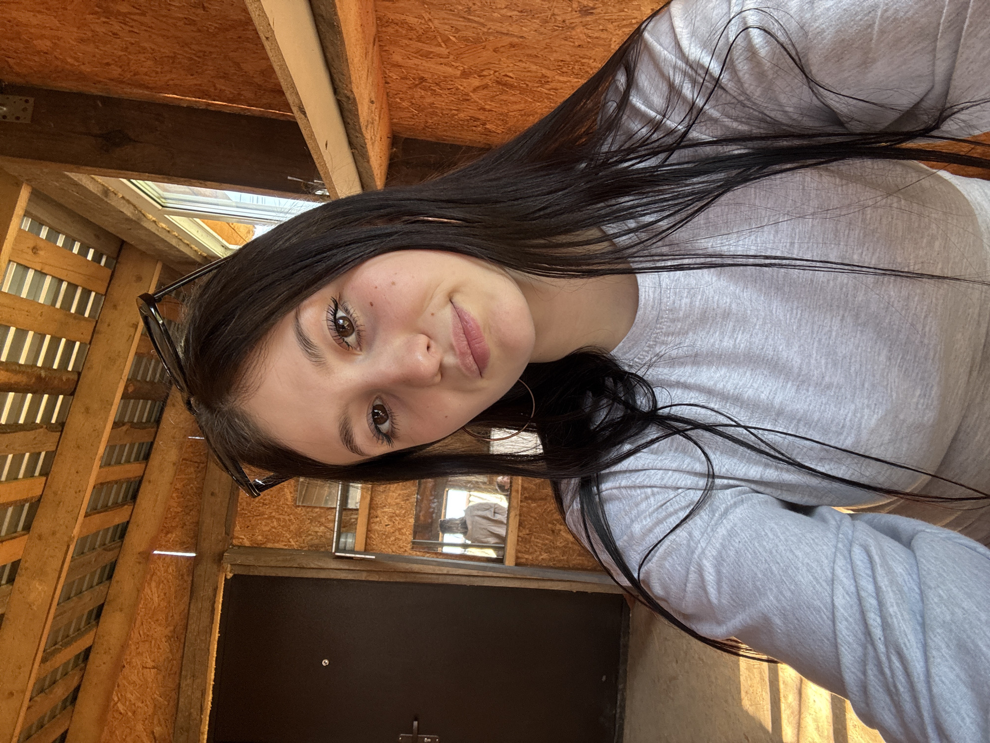 A young woman with dark hair and brown eyes poses for a selfie. She wears a light gray long-sleeved shirt and hoop earrings. Black glasses are perched on her head. The background shows an unfinished interior with exposed wooden beams and plywood walls. A black door is visible behind her. Natural light enters from a skylight. The photograph does not provide any indication of the city of Фоки, Russia.