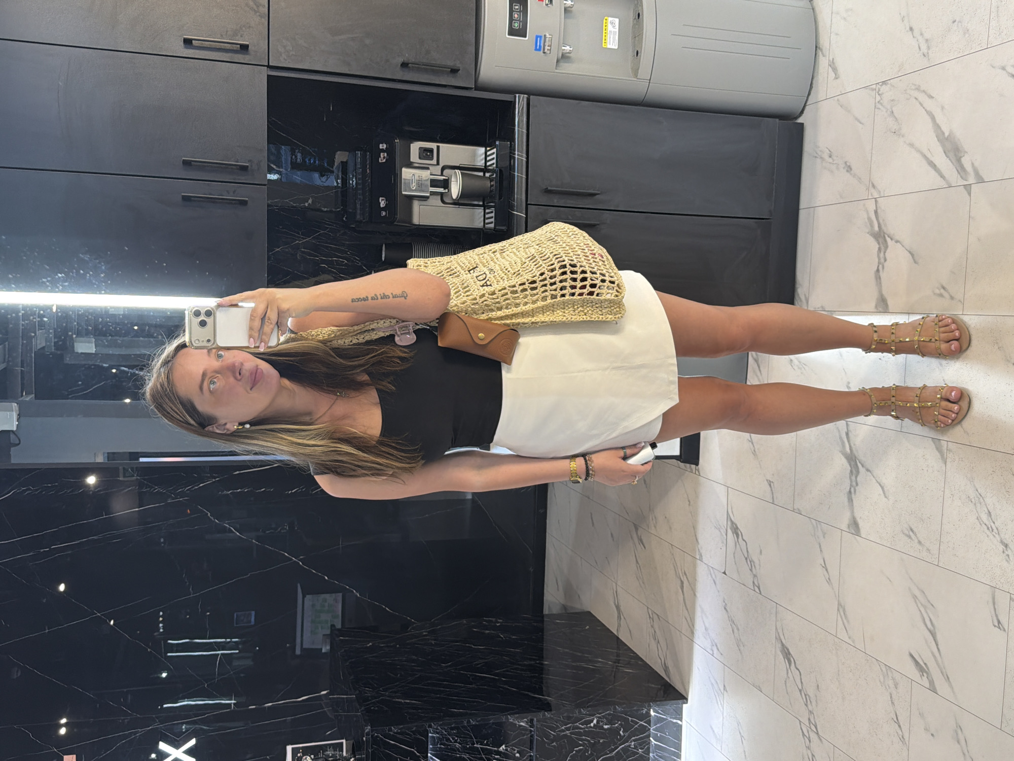 A woman stands in a brightly lit, modern room, taking a selfie in a mirror. She is wearing a black tank top, a white wrap skirt, and gold studded gladiator sandals. She has long, wavy brown hair and is wearing gold hoop earrings. A woven tote bag with a brown leather case attached hangs from her left shoulder. The background features black marble walls, dark cabinets, a coffee machine, and a water cooler. The floor is tiled in a light marble pattern. The location is the city of Укромновское сельское поселение, Ukraine.