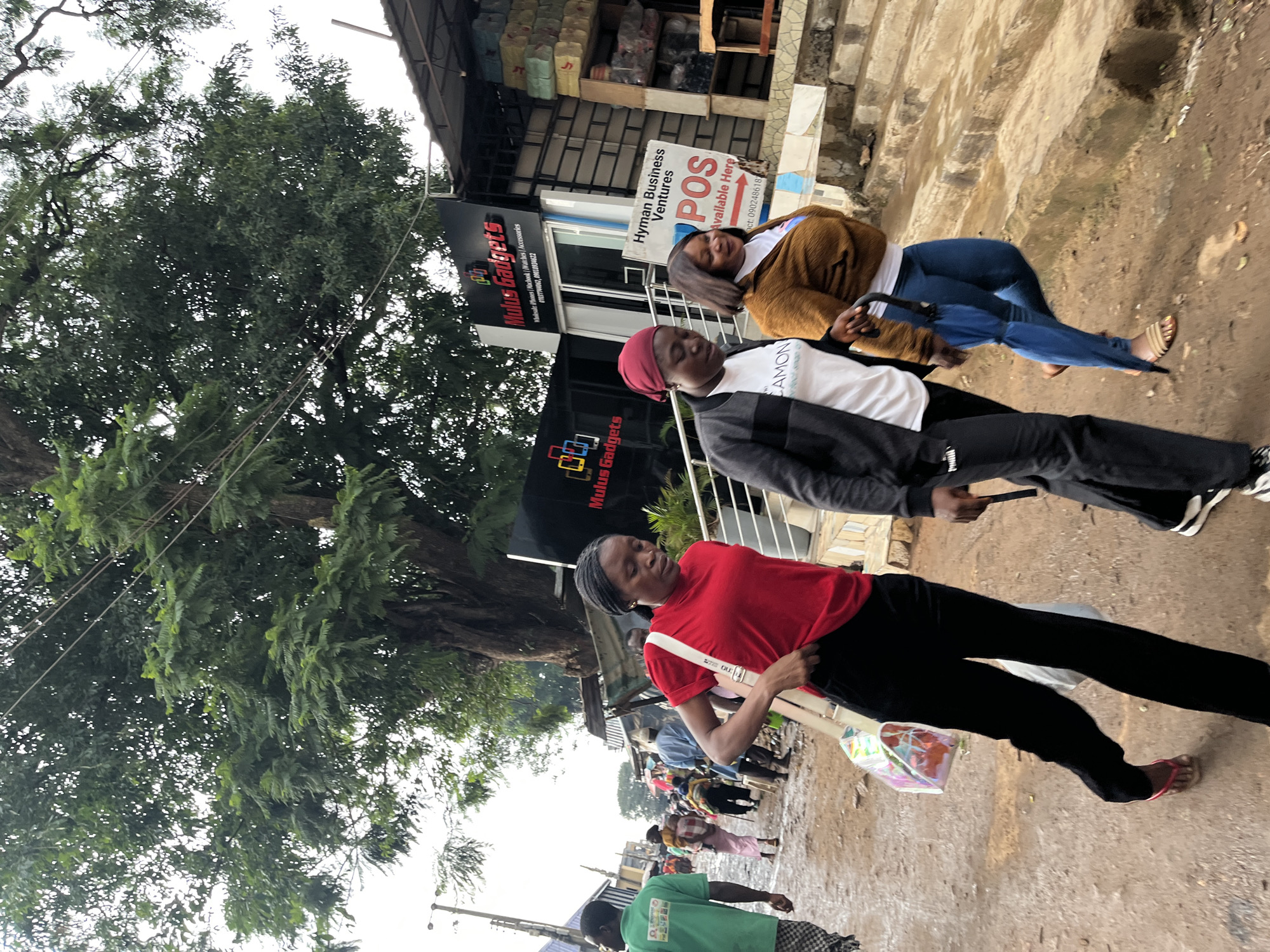 This is a street-level, eye-level, full-shot photograph taken in Jos, Nigeria. Three women are walking on a dirt and gravel path, some of it wet, suggesting it might have recently rained or is in a damp area. On the left, a woman in a red t-shirt and black pants walks with a shoulder bag. Next to her, a woman in a black jacket, white t-shirt, and black pants walks with her head down. To the right, a woman in a brown jacket and blue jeans walks with an umbrella and another person partially visible. Behind them, other pedestrians and market stalls line the street under a large tree with dense foliage. A sign for "Mulus Gadgets" is visible on a building in the background, along with another sign for "Hyman Business Ventures POS Available Here." The overall scene depicts a busy, informal commercial area.