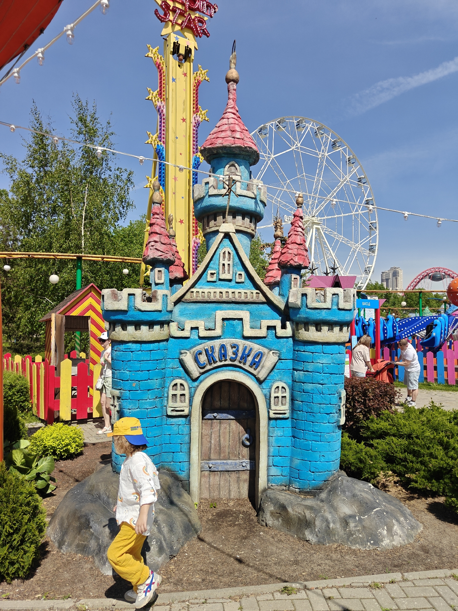 The image shows a child wearing a yellow hat and yellow pants walking past a small, blue castle-like structure. The structure has the word "СКАЗКА" written above the door. In the background, there's a Ferris wheel and a tower ride, suggesting an amusement park setting. Other people are visible in the background, near a colorful fence and what appears to be another ride. The scene is located in the city of Khimki, Russia, based on the presence of the "СКАЗКА" castle which is a recognizable landmark in the area. The weather is sunny, as indicated by the bright lighting and clear sky.