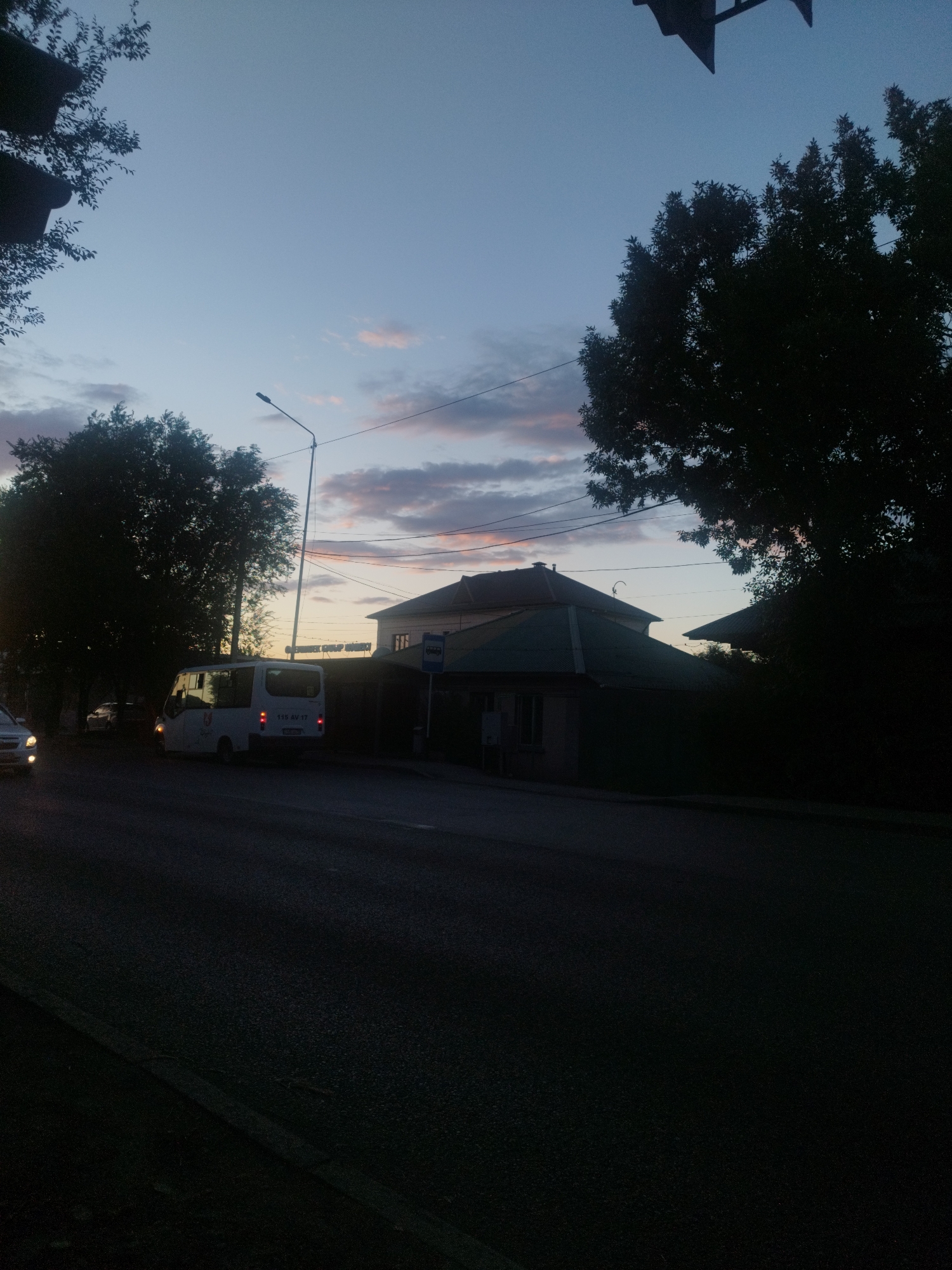 The image depicts a street scene in Shymkent, Kazakhstan, during twilight. The sky is a gradient of deep blue to lighter hues, with wispy clouds tinged with pink and orange. Silhouetted against the sky are the branches of large trees. In the middle ground, a white bus with its taillights illuminated is parked on the side of the road. Across the street, a building with a green roof and a bus stop sign is visible. The foreground consists of a dark asphalt road. The overall atmosphere is calm and subdued, characteristic of early evening.