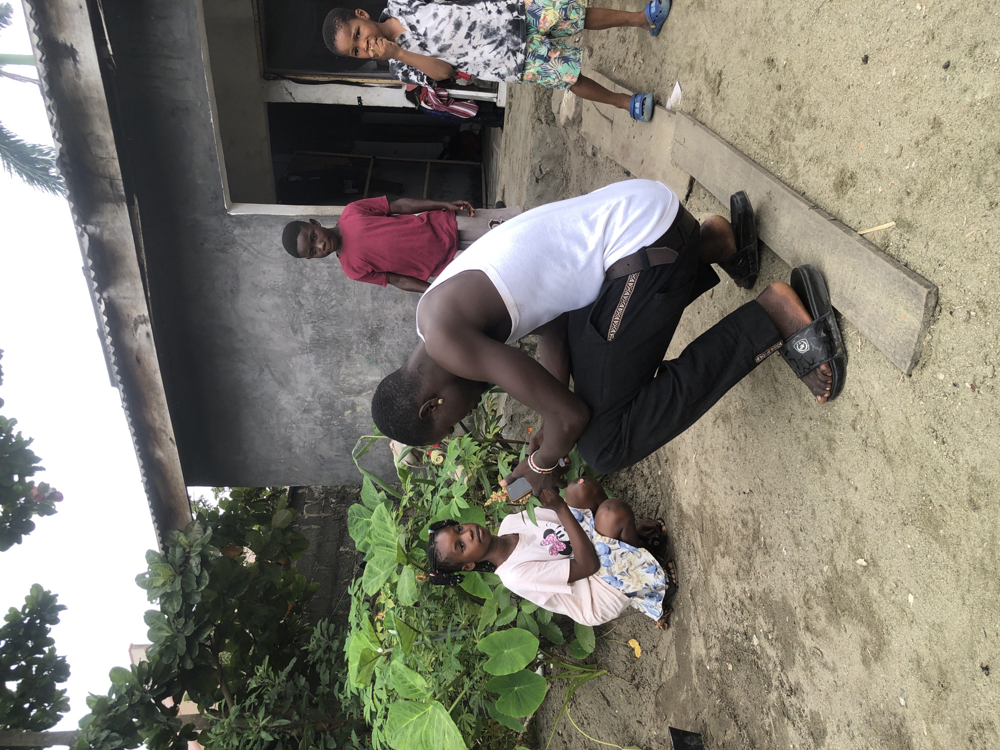 In Ado, Nigeria, a young man in a white tank top and black pants is crouching down, holding a mobile phone and appearing to interact with a young girl who is also crouching. The girl is wearing a light-colored t-shirt with a cartoon character and a patterned skirt. Behind them, two young boys are standing. One boy in a red t-shirt and shorts stands near a building, and another boy in a tie-dye t-shirt and patterned shorts is to the right. The environment is outdoors, with a rough, sandy ground and lush green foliage to the left. A simple building with a corrugated roof and grey concrete walls is visible in the background, suggesting a residential or informal settlement area. The lighting indicates it is daytime, with an overcast sky.