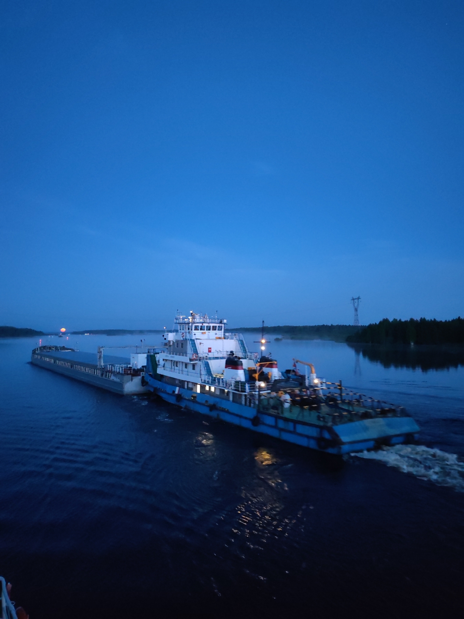 The image depicts a tugboat pushing a barge on a river, possibly the Volga River, at dusk. The scene is possibly near Chagino, Russia, though without definitive landmarks it's difficult to confirm. The tugboat is light blue and white with a multi-tiered structure and lights visible. It's pushing a dark grey barge. The river is dark with reflections. In the distance, there's a faint orange glow on the horizon, possibly from the setting sun. On the riverbank, there is a forest. A power line tower is seen on the right bank. There are no people visible. The primary action is the movement of the tugboat and barge through the water.