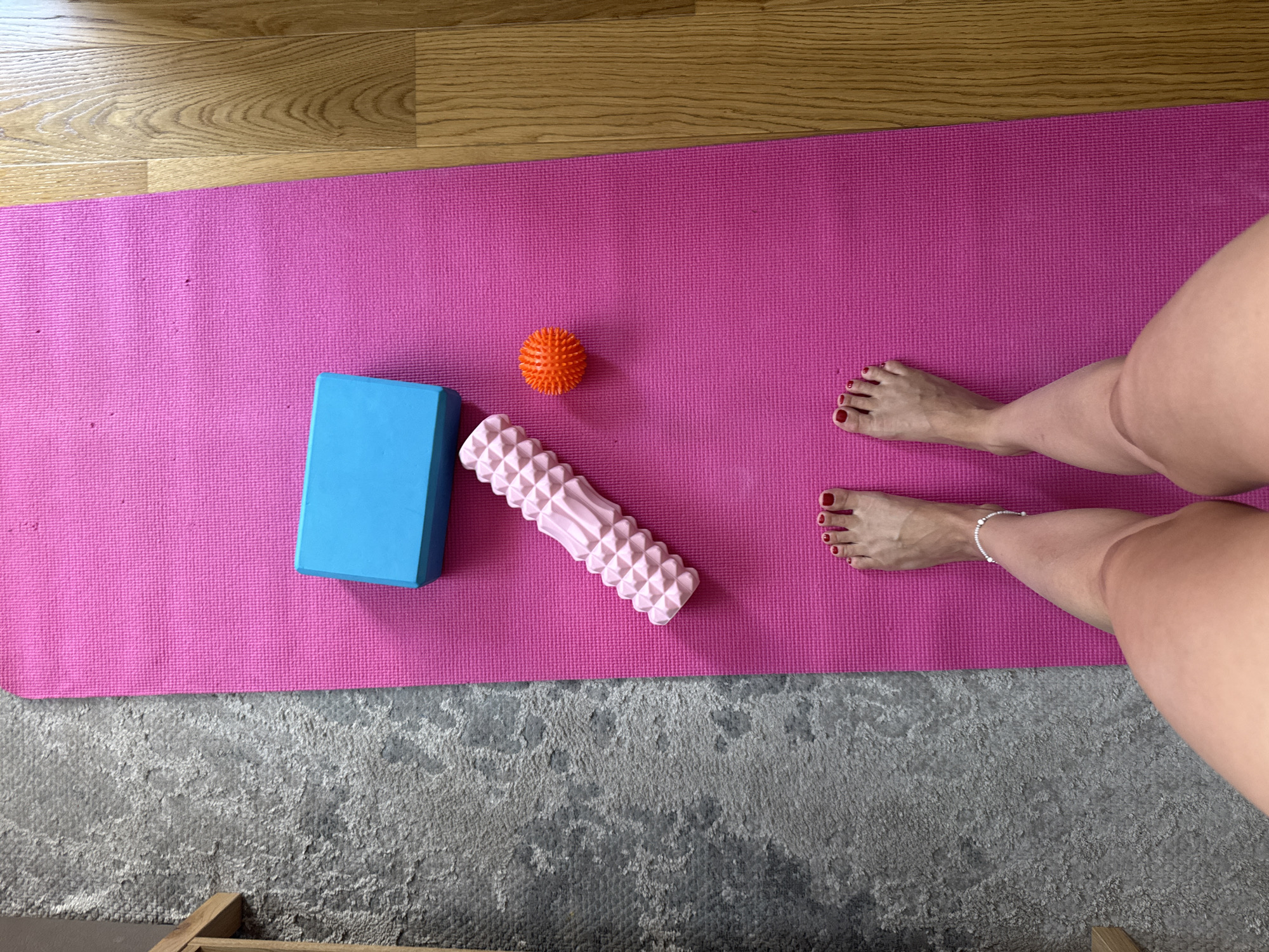 The image shows a top-down view of a pink yoga mat laid out on a wooden floor. On the mat are several items: a bright blue yoga block, a pink textured foam roller, and a small orange massage ball. To the right of these items, a person's legs and feet are visible, positioned on the yoga mat. The person has red nail polish on their toenails and is wearing a delicate silver anklet. The scene suggests an individual is preparing for or has just finished a yoga or physical therapy session. The lighting appears to be natural, indicating it could be daytime. The setting is a home or studio environment, with a rug visible below the yoga mat and part of a wooden furniture piece in the lower left corner. There is no visible text in the image.