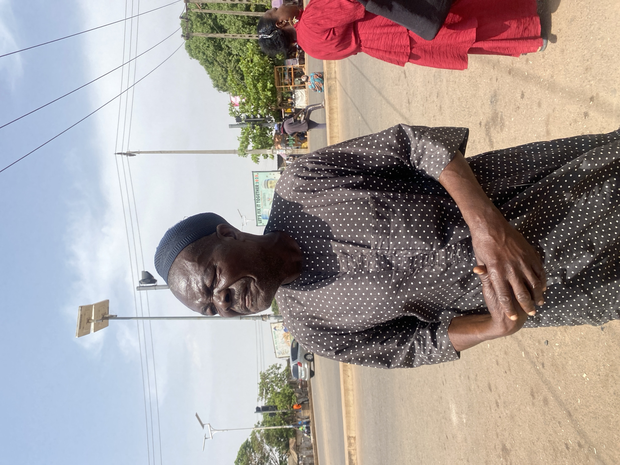The image captures a sunlit street scene in Sabon Tasha, Nigeria. In the foreground, an elderly Black male is positioned slightly left of center, visible from the chest up. He wears a dark knitted cap and a dark long-sleeved shirt adorned with white polka dots. His face is turned partially to the right, with his eyes squinting, and his hands are clasped together at waist level. To the right, a female is partially visible, wearing a bright red tiered dress and black shoes, with a white accessory in her dark hair.
The immediate ground is an asphalt road. In the mid-ground, a wider asphalt road is present, featuring a concrete median. A silver SUV is observed on the left lane. Overhead, a clear sky with scattered clouds is visible. Multiple utility poles line the street, some fitted with solar panels and streetlights, and others supporting traffic signals and overhead power lines. A billboard displaying the text "LET'S FIX IT TOGETHER 2023." and an image of a man is situated behind the road. Green foliage from trees is visible on the right side of the frame. In the background, further down the road, indistinct figures are visible, including one person in a dark shirt and blue cap, and another seated on the ground, suggesting market activity or street vendors near rudimentary structures.