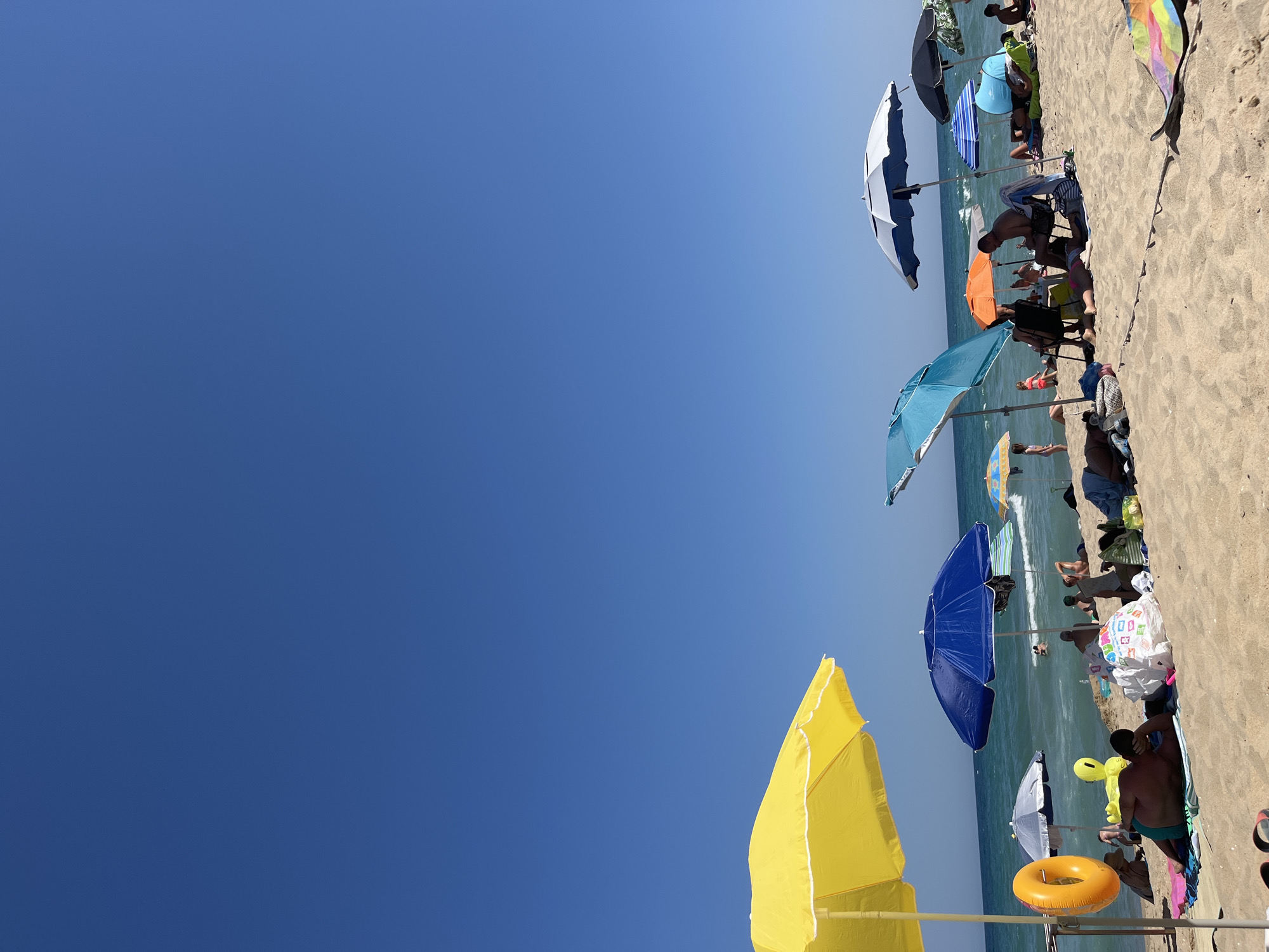 A wide shot of a crowded beach on a sunny day shows numerous beach umbrellas and people relaxing on the sand. The ocean is visible in the background with moderate waves, and swimmers can be seen in the water. In the foreground, a yellow umbrella stands out, with an orange inflatable ring hanging from its pole. Several individuals are lying on towels and sunbathing. People are seated under various umbrellas, some engaging in conversation, while others are observing the sea. The sand is light brown, and the sky is a clear, deep blue. This scene is a typical summer day at the beach in Obzor, Bulgaria.
