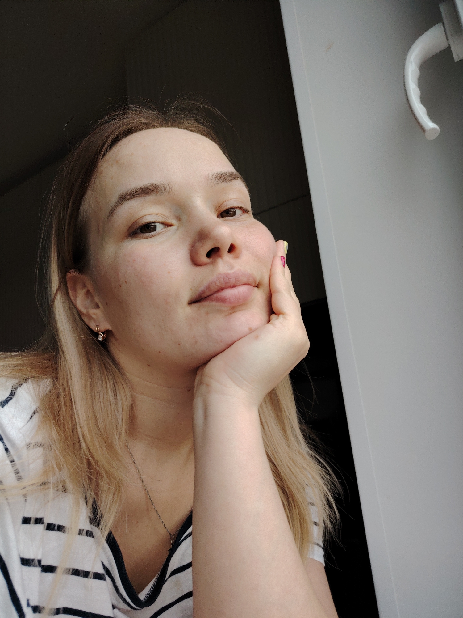 A young woman is seated indoors, possibly near a window, as indicated by the natural lighting. She is resting her face on her hand, her expression thoughtful. The woman has fair skin, light brown hair, and brown eyes. She is wearing a striped T-shirt and a delicate necklace. The environment consists of a white door frame and adjacent dark space, providing a stark contrast with the brightly lit area where the woman is situated. The location is not specified by any visual cues within the image. Марковский, Russia cannot be confirmed.