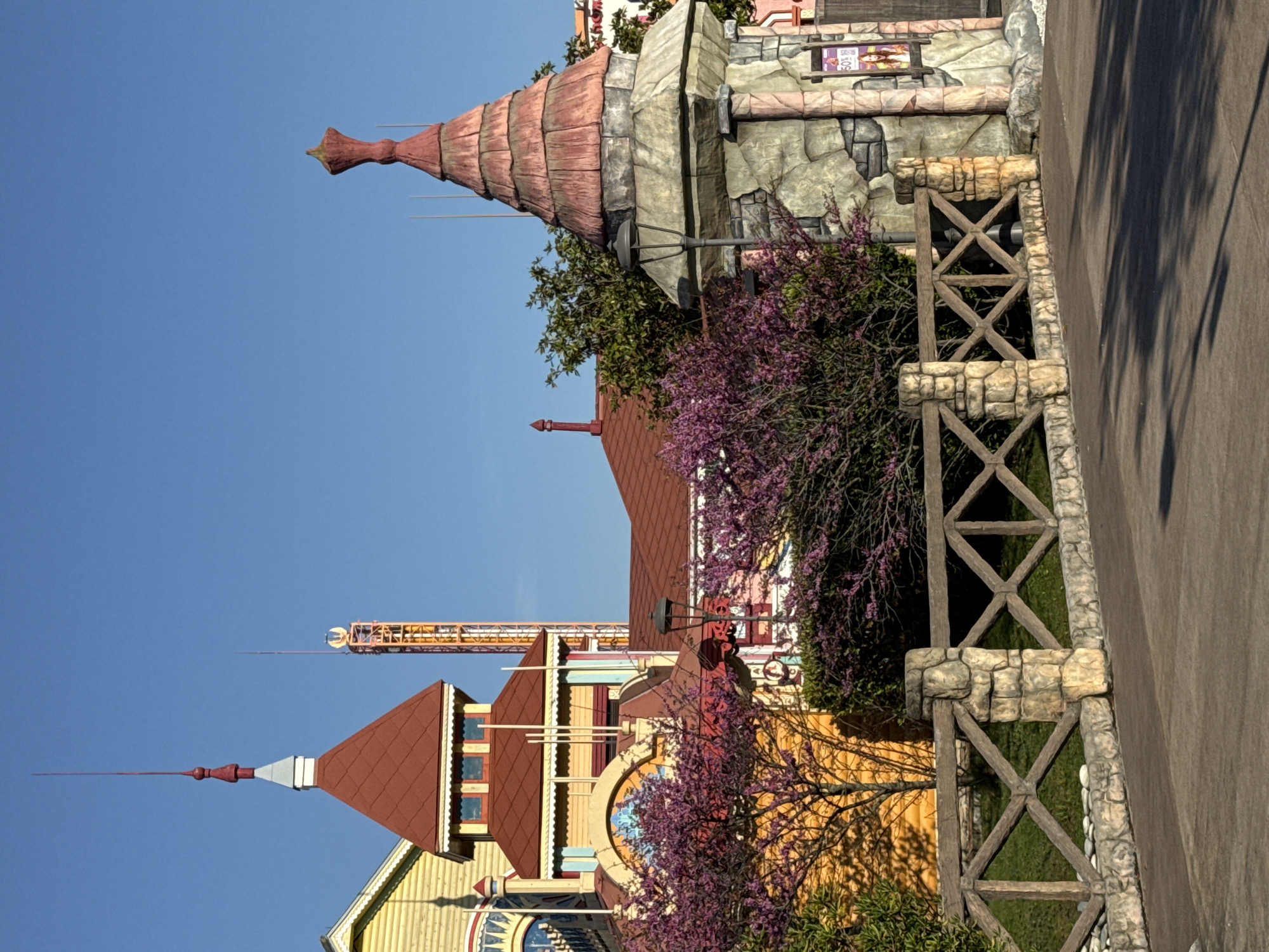 The image captures a scene in Sochi, Russia, featuring a whimsical collection of architectural elements. A building with a tall, conical tower resembling a wizard's hat dominates the upper portion. Below this, a complex of buildings with peaked roofs, turrets, and decorative facades creates a fairytale ambiance. A tall, orange-colored structure can be seen in the background, possibly a ride or attraction, adding to the amusement park atmosphere. A rustic fence with stone and wooden supports runs along the right side of the frame, separating the buildings from a paved walkway. Trees with vibrant purple blossoms add a touch of natural beauty to the scene. The sky is clear blue, suggesting a bright, sunny day.