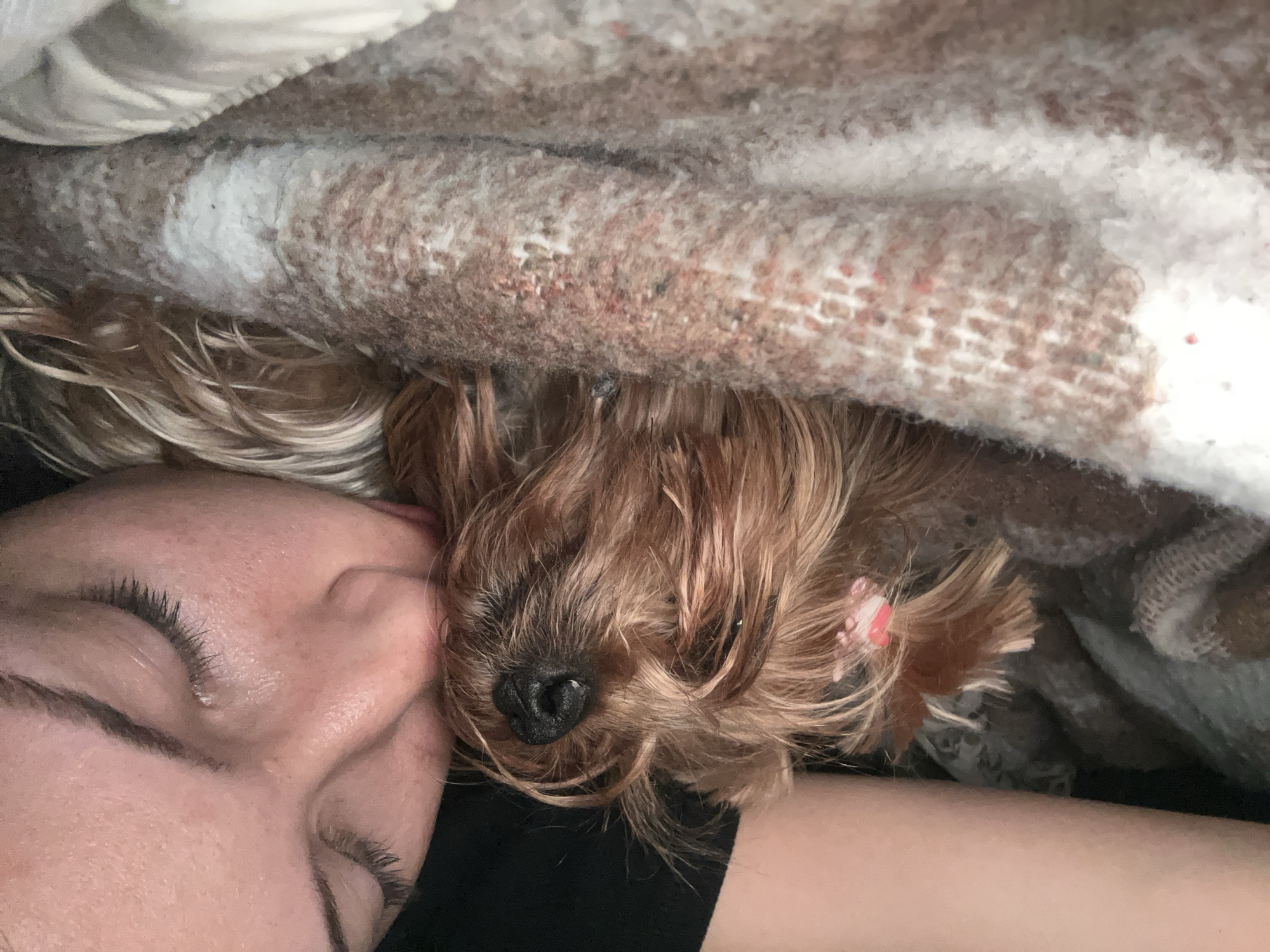 A person is lying in bed with a small brown dog. Both appear to be sleeping. A patterned blanket covers them. The person is fair-skinned with closed eyes and long eyelashes. The dog has long, wavy, brown fur. The dog is wearing a small pink bow or clip on its head. The person is wearing a black top. The photo is taken from above. There is no indication of the location being Burgas, Bulgaria, or any other geographical information.