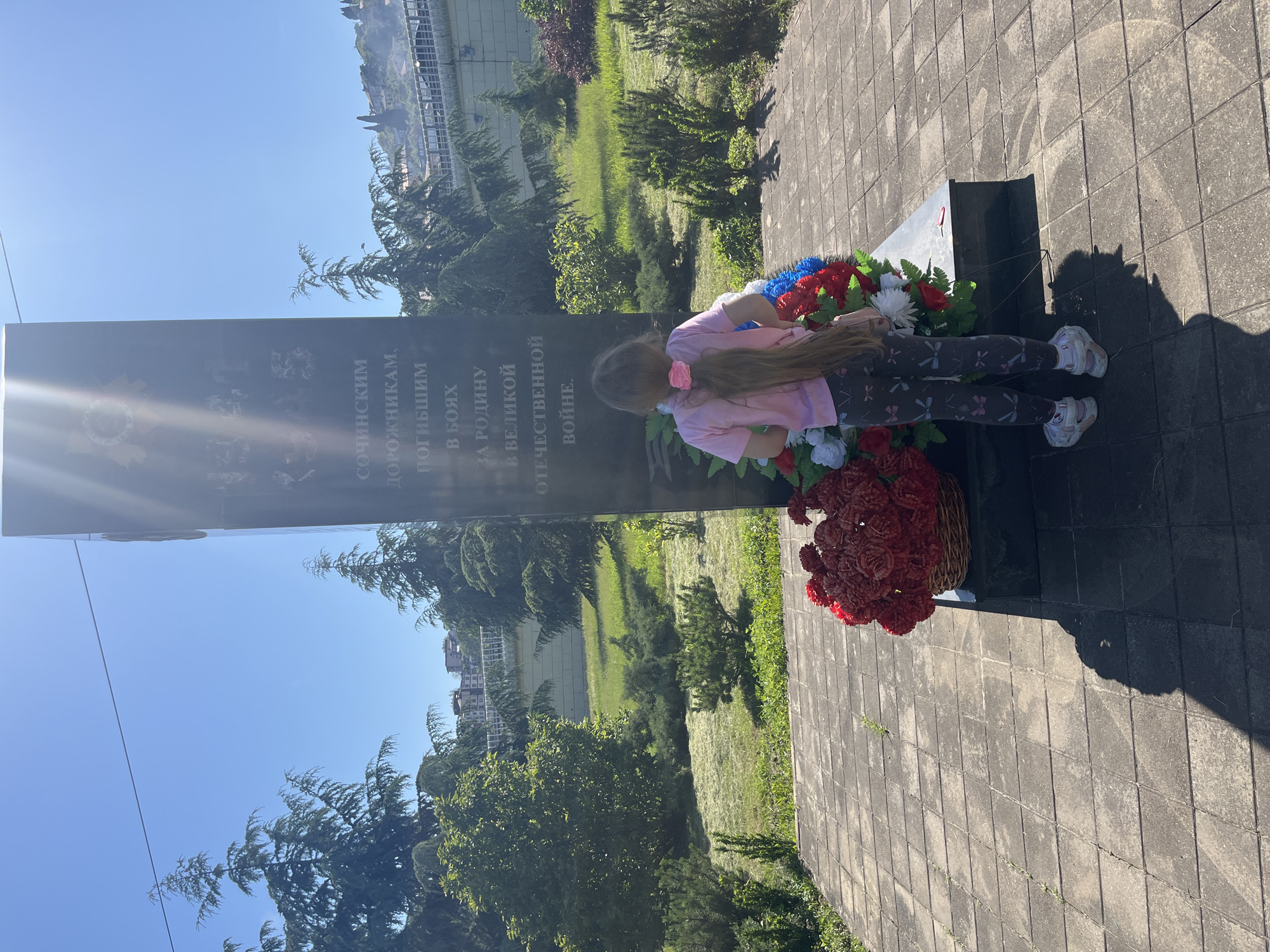 The image, oriented horizontally, depicts a young girl at a war memorial under a clear, sunny sky in what appears to be Sochi, Russia. The girl, with long, light brown hair secured by a pink bow, wears a pink t-shirt, dark patterned leggings, and white sneakers. She is bending over towards the base of a tall, dark, rectangular monument, which is adorned with several floral arrangements.
In front of the monument, there is a large wreath containing red, white, and blue artificial flowers, reminiscent of the Russian flag's colors. To the right of this wreath, a wicker basket overflows with numerous red artificial flowers, likely carnations or roses. The girl's posture suggests an act of contemplation or placing flowers in remembrance.
The monument itself features Cyrillic text, partially visible and slightly obscured by glare. The discernible text reads:
"СОЧИНСКИМ ДОРОЖНИКАМ, ПОГИБШИМ В БОЯХ ЗА РОДИНУ В ВЕЛИКОЙ ОТЕЧЕСТВЕННОЙ ВОЙНЕ."
This translates to: "To the Sochi road workers, who fell in battles for the Motherland in the Great Patriotic War."
The setting is an outdoor area, likely a park or a memorial square. The ground is paved with grey interlocking tiles. Beyond the monument and the paved area, there is a grassy expanse with scattered trees and bushes. In the background, a distant building or structure can be seen. The bright sunlight casts distinct shadows on the paved ground, indicating mid-day. The overall scene conveys a solemn act of remembrance and respect for those who died in the Great Patriotic War.