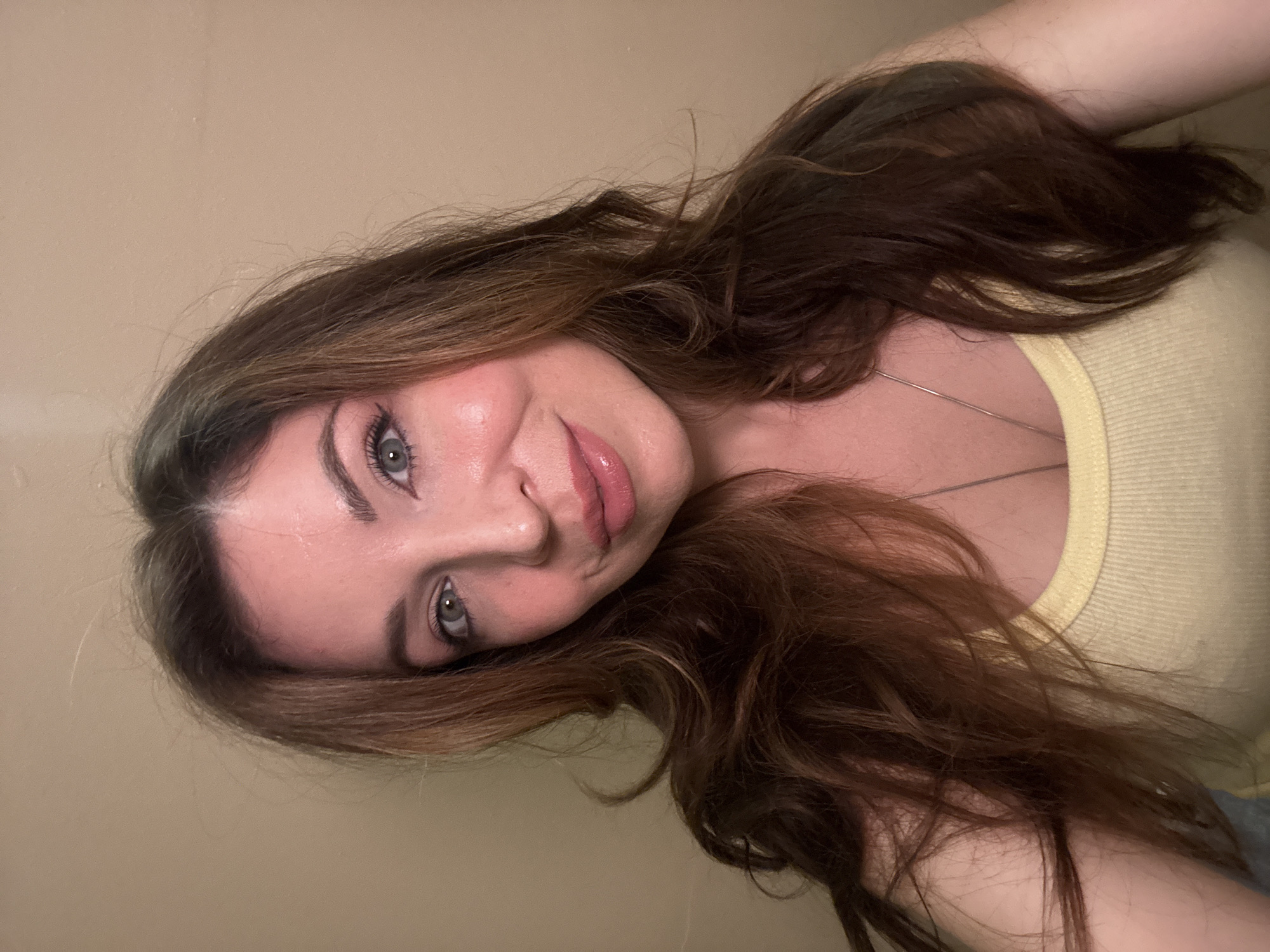 This is a close-up selfie of a Caucasian woman with medium-length, wavy brown hair. She is looking directly at the camera with a slight smile. She is wearing a yellow ribbed tank top and a silver necklace. The background is a plain, light-colored wall. The scene is indoors, with soft lighting. The image was taken in Odesa, Ukraine.