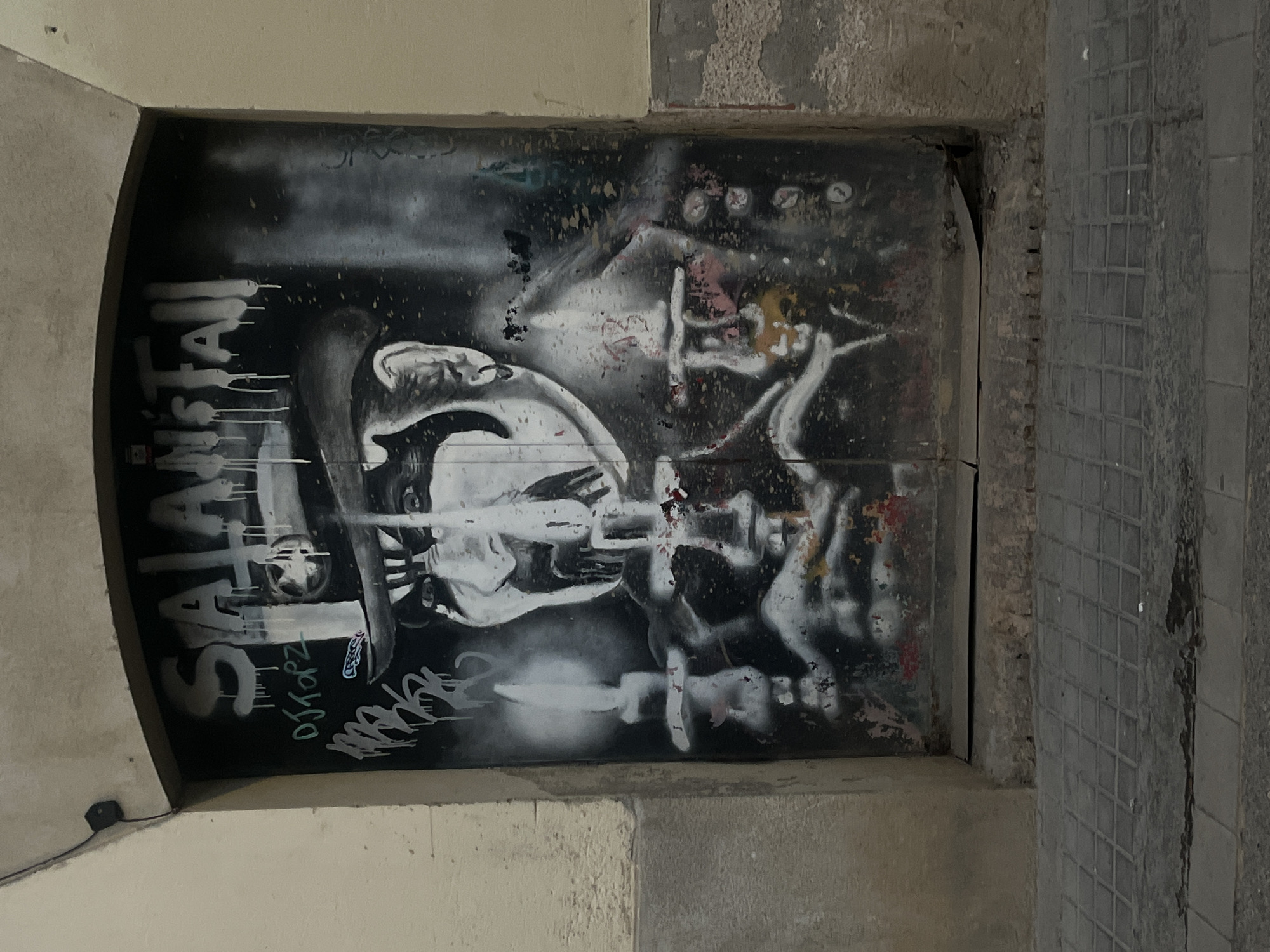 A low-angle, medium shot depicts a section of graffiti-covered wall in what is identified as Barcelona, Spain. The graffiti, rendered primarily in black and white with some red and yellow, fills a rectangular frame, possibly a doorway or alcove. The image is an abstract composition of figures, forms, and the word "SATANSTAI" vertically oriented on the left side. Beneath the name “SATANSTAI” the graffitti reads “DJ TOOPZ”. The surrounding wall is weathered, with varying textures and tones of gray, suggesting age and exposure. The scene is dimly lit, lending a somber or mysterious atmosphere. To the right of the graffiti, tiles can be seen along the outside of the wall. The bottom and left side walls are light colored and smooth.