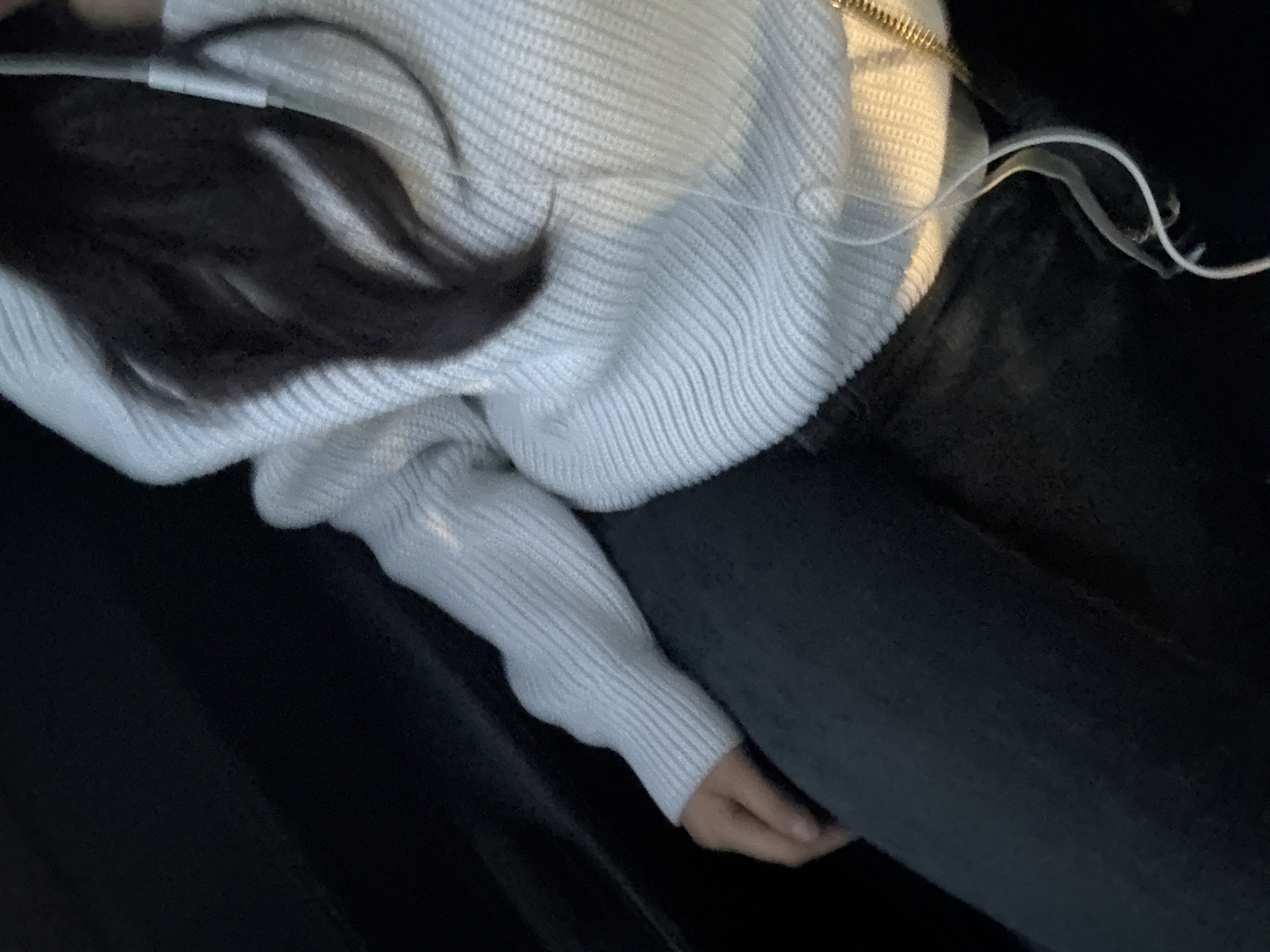 A person is seated, wearing a white ribbed knit sweater and dark jeans. The person has dark hair. White headphone wires are visible. The location is unknown and there is no information to suggest it is in Puebla, Mexico.