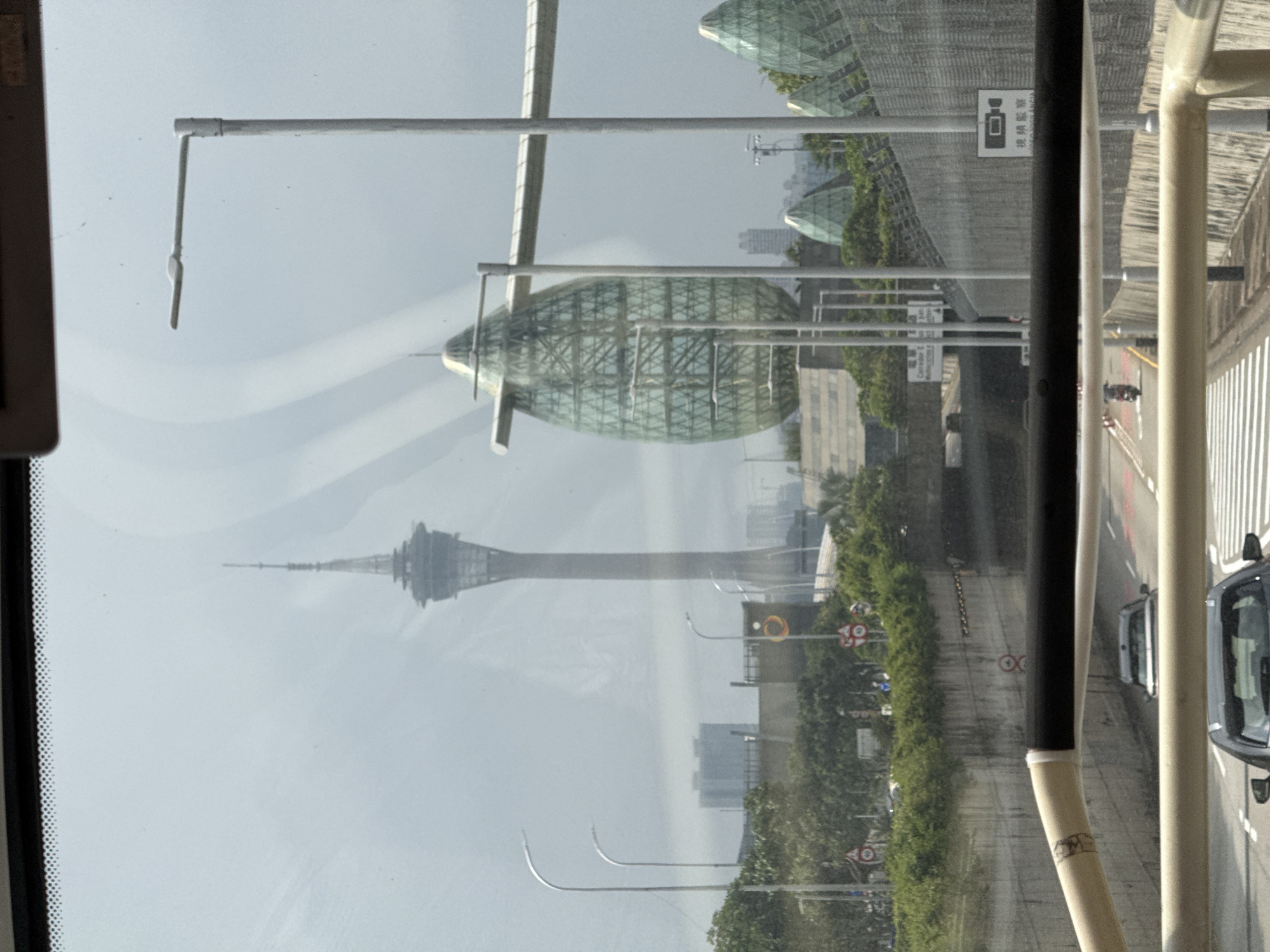 The image captures a view from inside a vehicle, likely a bus or car, showcasing a blurred cityscape of Unknown, Macao. Through the window, the prominent Macau Tower stands tall, while the Science Center’s geometrically shaped buildings are also visible. The scene is oriented vertically. A road with traffic including at least one car is seen to the right. Various street lamps and signs line the road. The foreground includes parts of the vehicle's interior, like the frame of the window and support structures. The overall atmosphere is hazy.