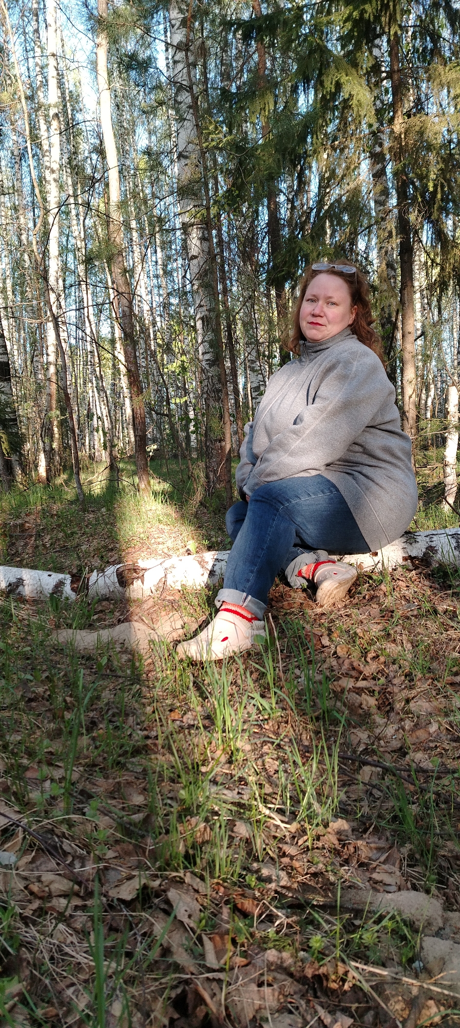 The photograph was taken outdoors in what appears to be a forest or wooded area in Zipunovo, Russia. A woman with reddish hair and glasses is sitting on a fallen birch log. She is wearing a gray jacket, blue jeans, and white sneakers with red trim. The ground is covered with grass and fallen leaves. Birch trees are visible in the background.