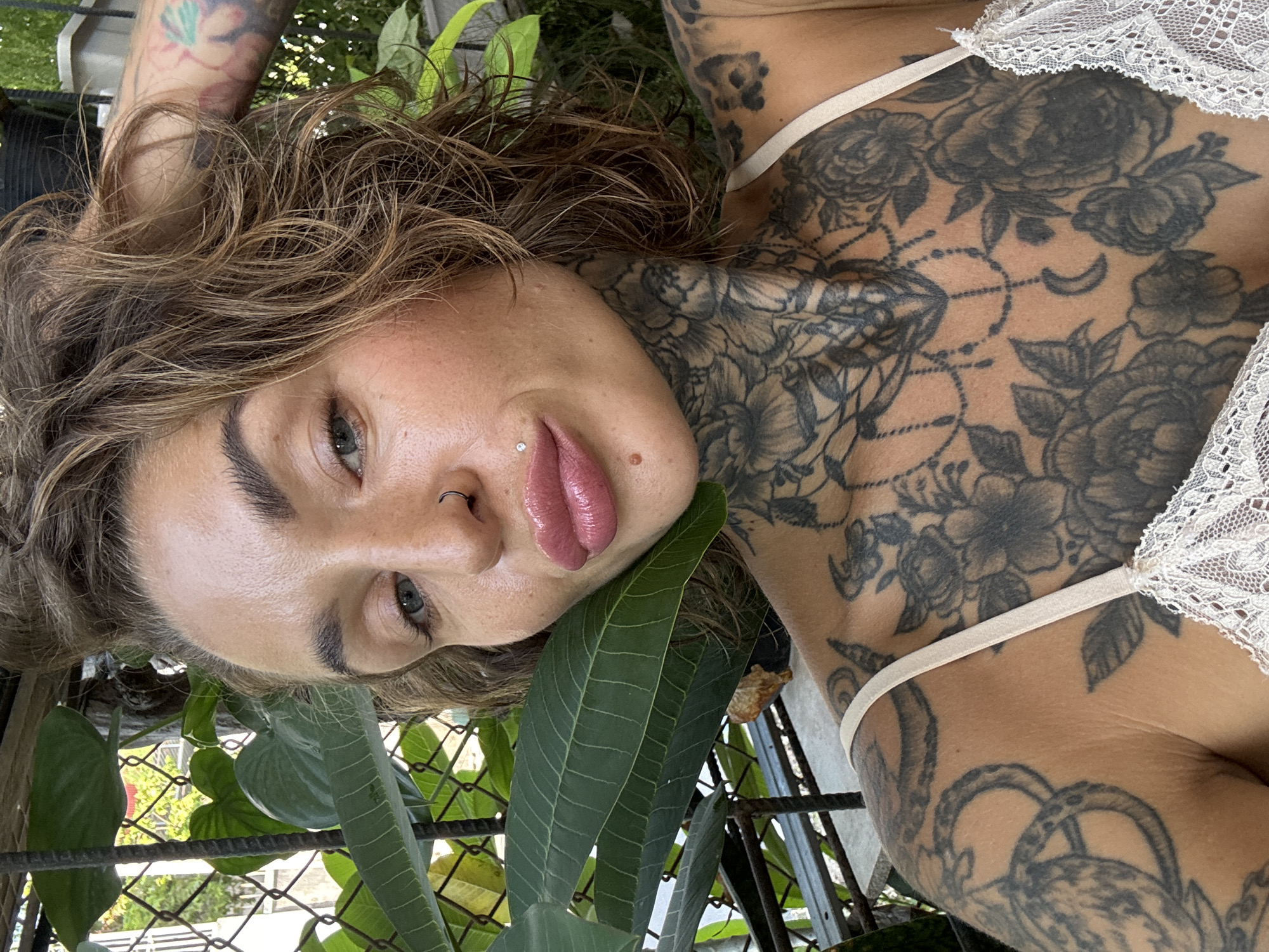 A woman with dark brown, wavy hair and intricate tattoos on her neck and chest is pictured in a close-up shot. She is wearing a light-colored lace top. Her eyes are open and she is looking towards the camera. The background consists of green foliage and a metal fence, suggesting an outdoor setting. The lighting is natural and appears to be daylight.