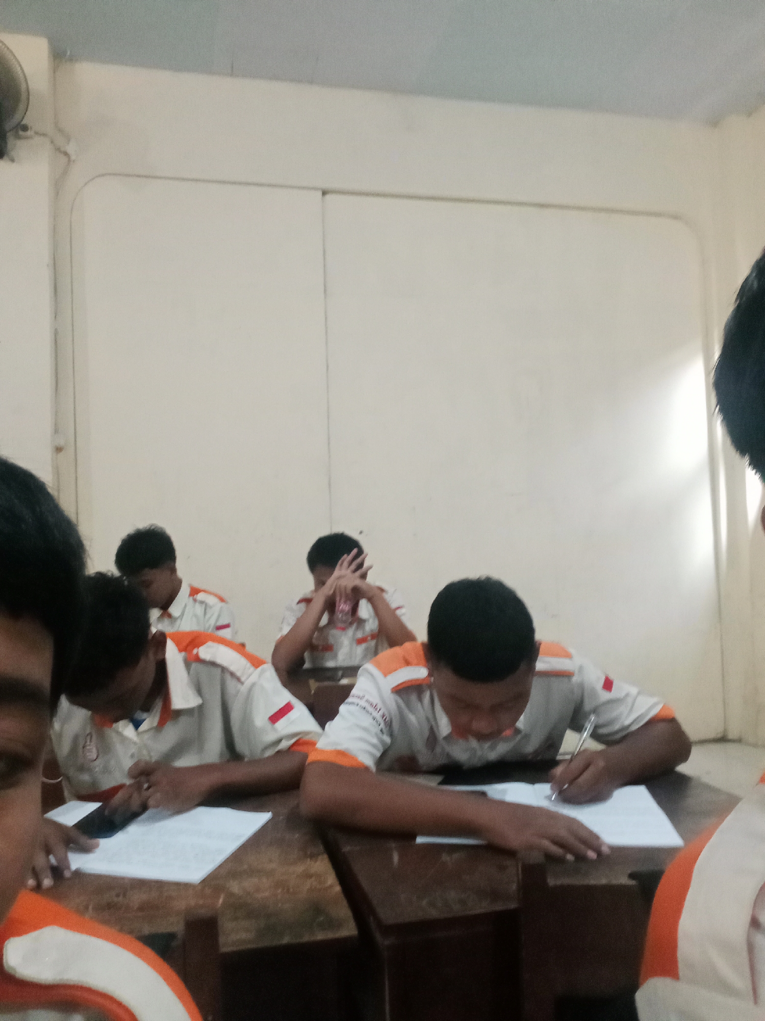 The image depicts a classroom scene in Secang, Indonesia, where several young men, presumably students, are seated at desks. They are all wearing similar white shirts with orange accents and red insignia, suggesting a school uniform. The students appear to be engaged in writing or taking notes on paper. The classroom walls are plain and light-colored, and the lighting suggests an indoor environment, possibly illuminated by artificial light or natural light from an unseen source. The overall atmosphere seems focused and studious, with some students looking down at their work and others with their hands on their heads, perhaps in thought or concentration. The desks are simple wooden structures.