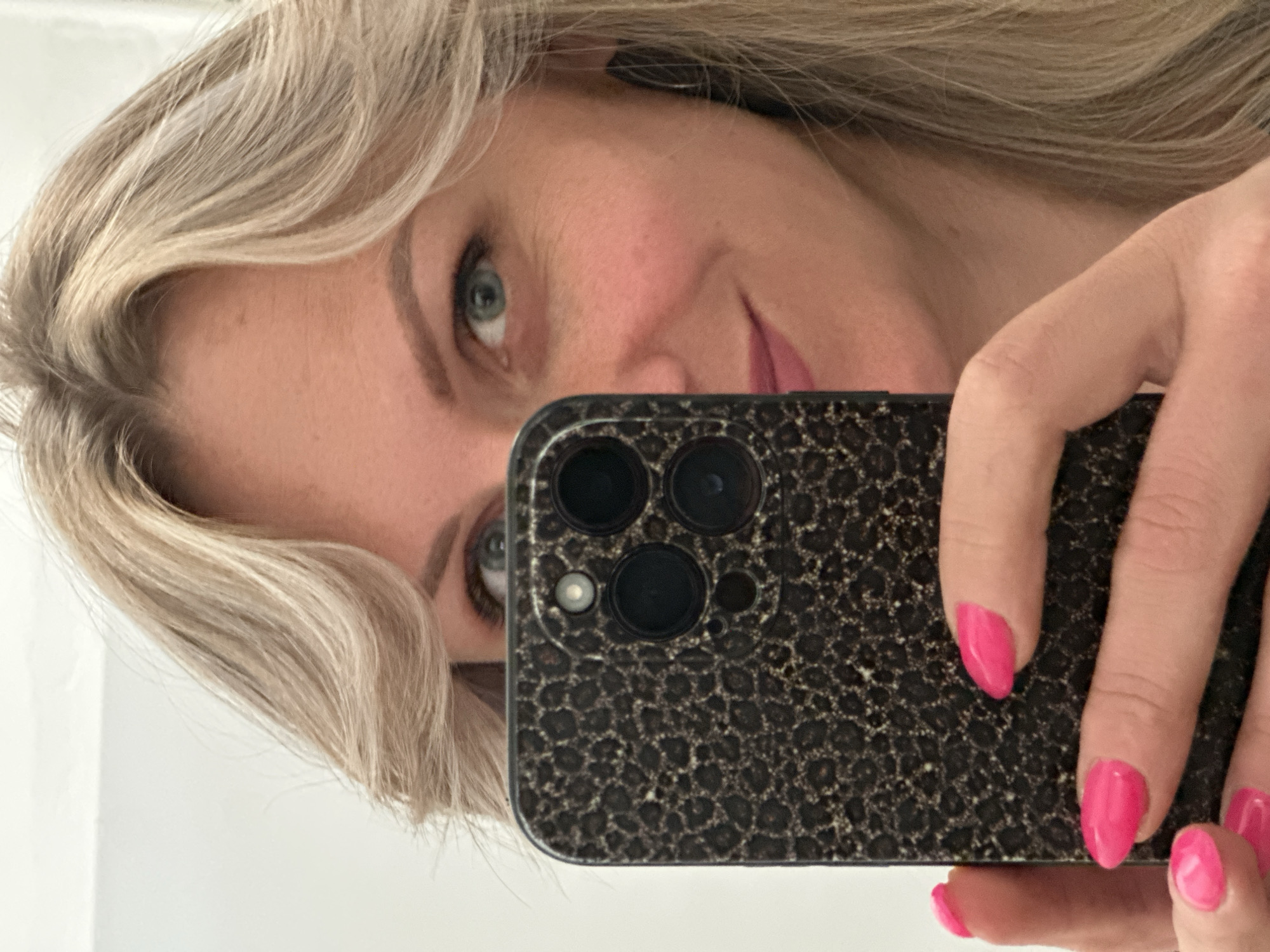 A close-up, rotated portrait captures a woman taking a mirror selfie indoors. Her fair-skinned face is partially visible on the left, framed by blonde or platinum hair styled with gentle waves. She has striking blue-green eyes, looking directly into the camera, and a subtle smile on her pink-lipsticked lips. Her eyebrows appear neatly groomed.
She holds a black smartphone in her right hand, which occupies a significant portion of the right side of the frame. The phone case features a textured, possibly animal print, pattern. The multiple camera lenses and flash unit on the back of the phone are clearly visible. Her fingernails are neatly manicured and painted a vibrant, glossy hot pink, providing a bright color contrast to the dark phone.
The background is a plain white surface, suggesting an interior wall. The lighting is bright and even. The image conveys a casual and pleasant moment. No text is visible within the image. The provided context indicates the location as Venafro, Italy.